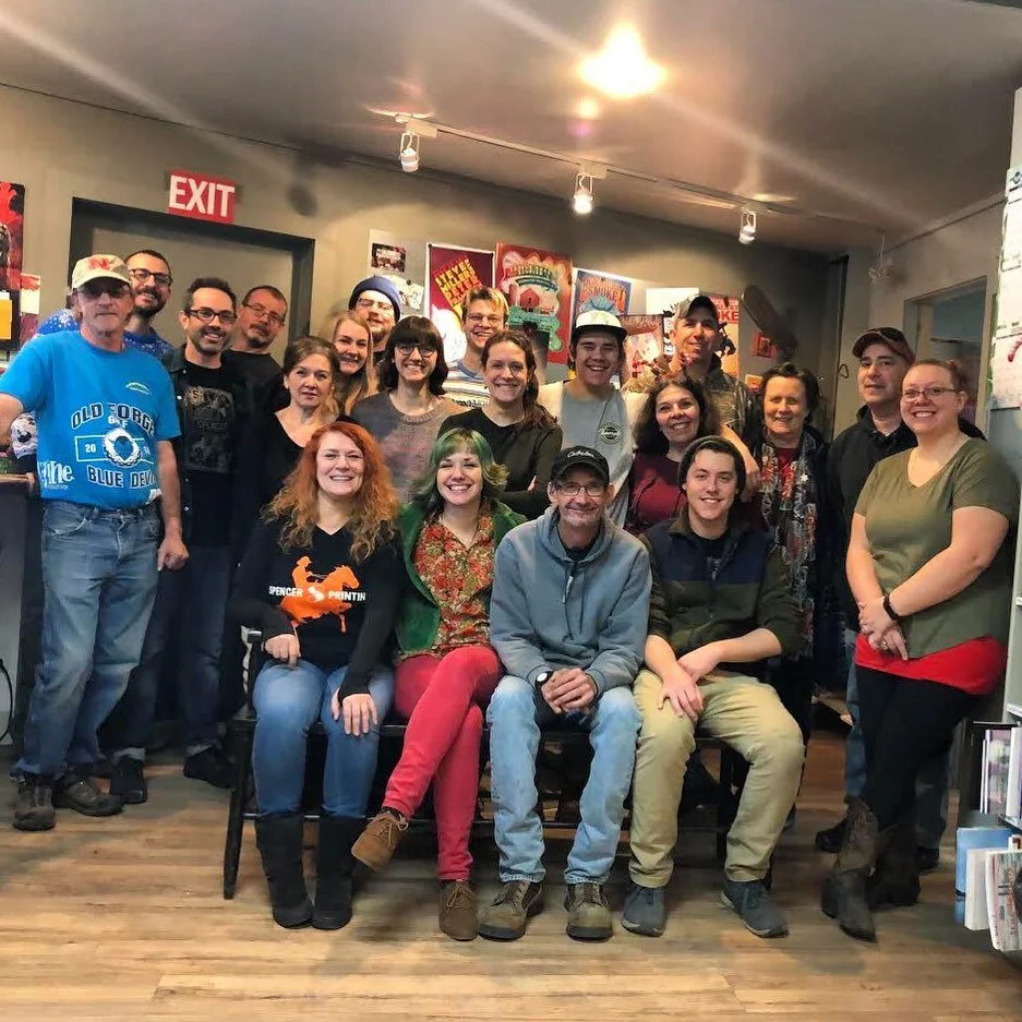 A group of 17 people posing together in a cozy indoor space, some sitting and some standing, with posters on the wall behind them and an exit sign above a door.
