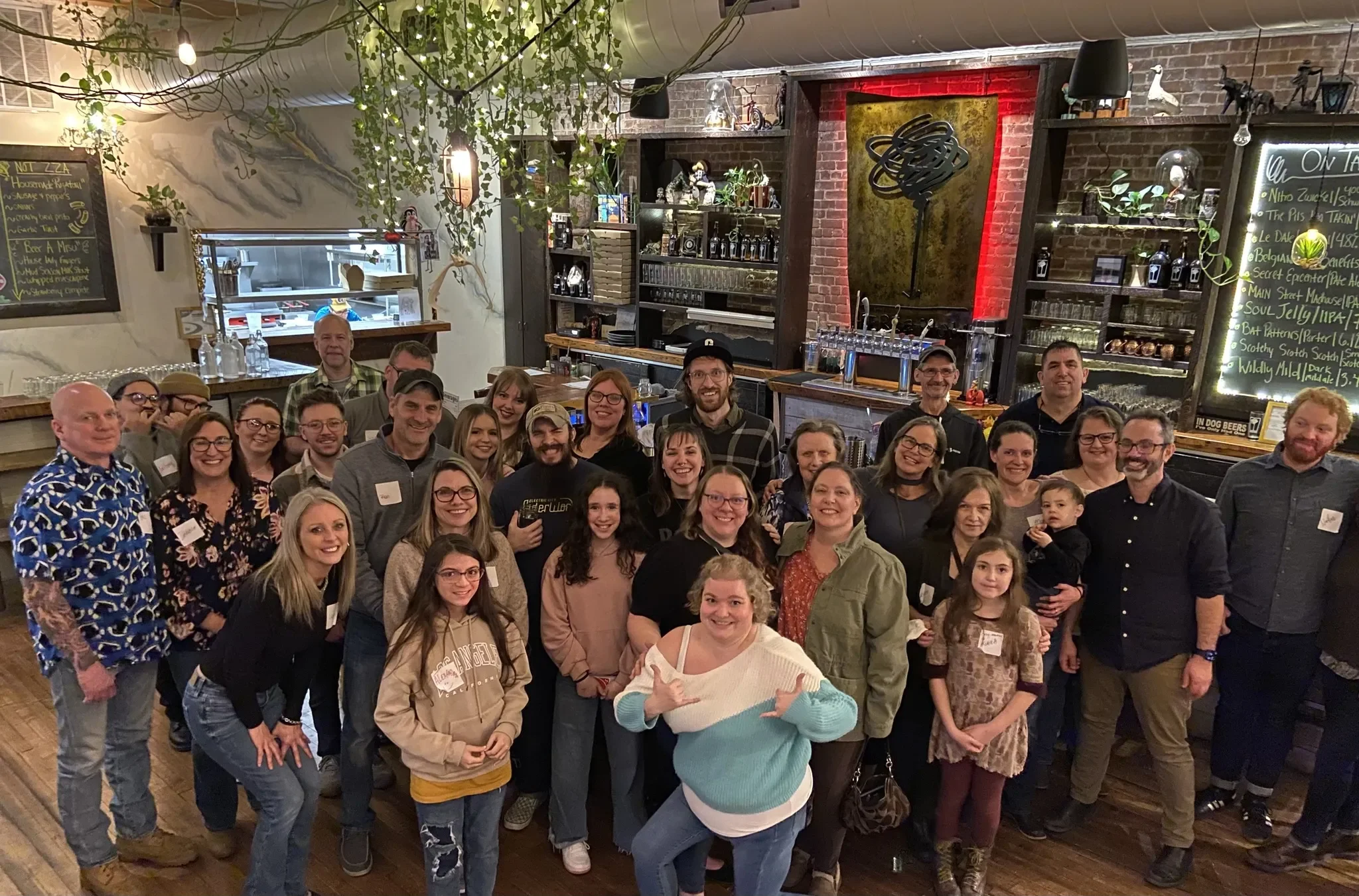 Group of smiling people posing for a photo in a restaurant or bar with exposed brick walls and decorative plants.