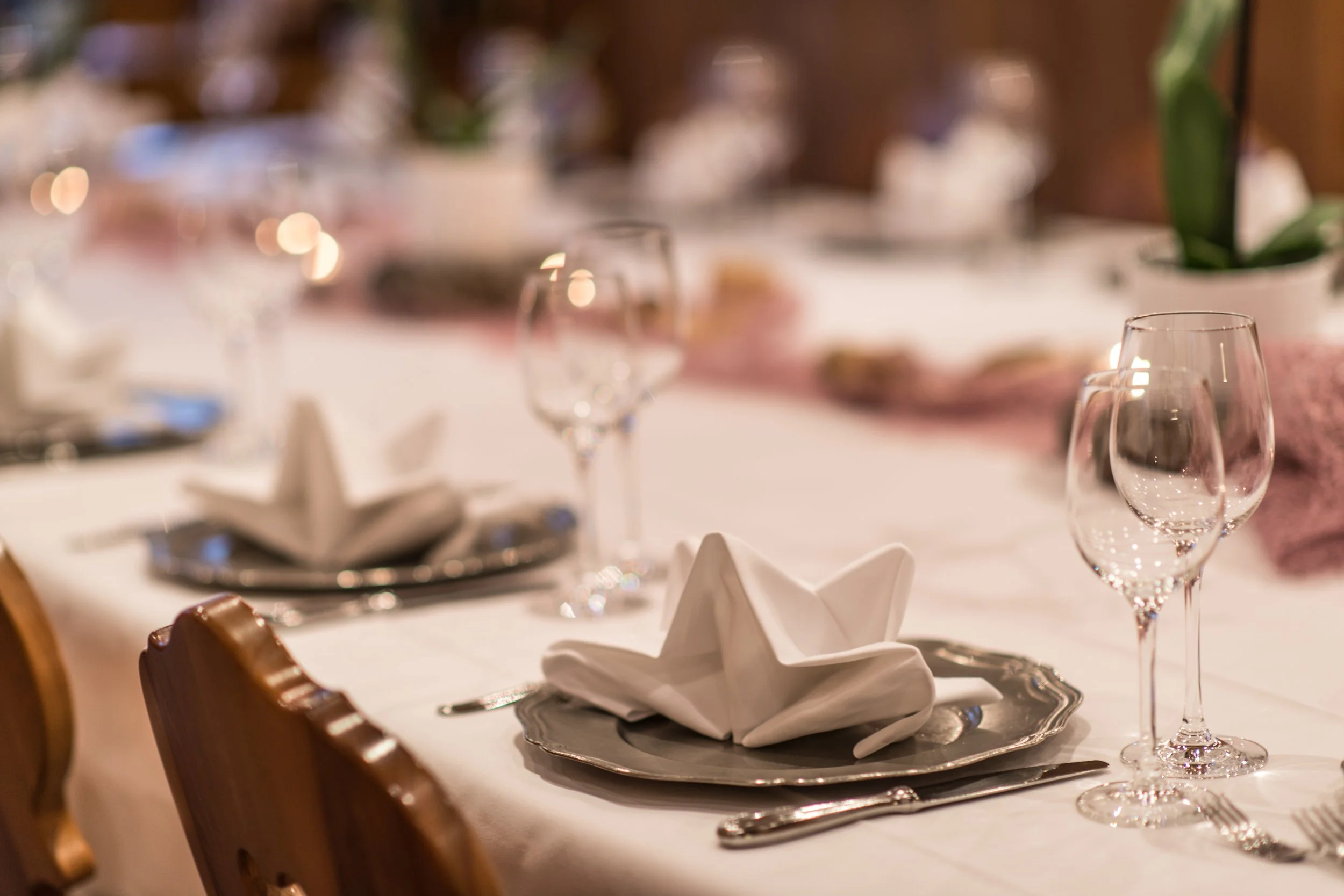 Ein festlich gedeckter Tisch mit Weingläsern, einer silbernen Servierplatte, weißen Stoffservietten, einer silbernen serving Platte und Tassen, im Hintergrund sind Blumen und Kerzen.