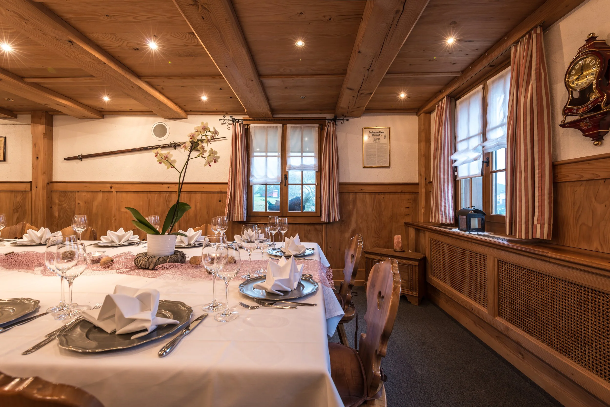 Ein festlich gedeckter Tisch in einem rustikalen Esszimmer mit Holzdecken, Fenstern mit weißen Vorhängen, einem Blumenarrangement und Kerzenleuchtern.