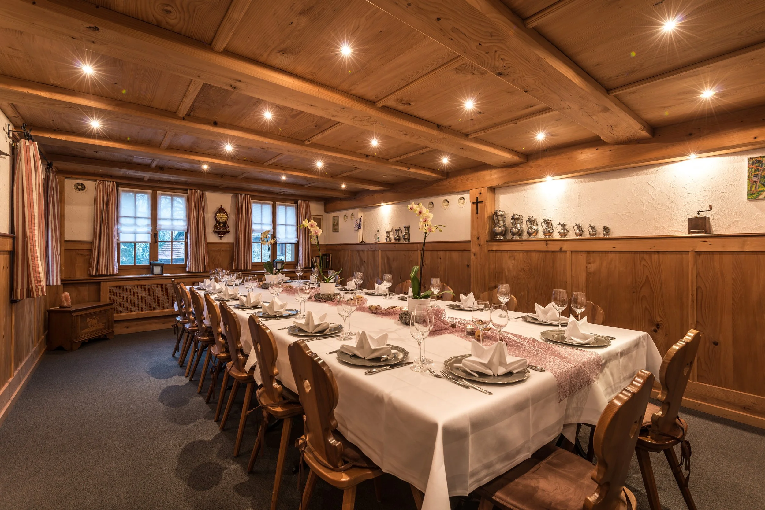 Ein festlich gedeckter Essbereich in einem rustikalen Raum mit Holzwandpaneelen, einem langen Tisch mit weißen Tischtüchern, Kerzen, Wein- und Wassergläsern sowie Organgematten. An der Wand hängen mehrere Silberkrüge und ein Kreuz, die Fenster sind m