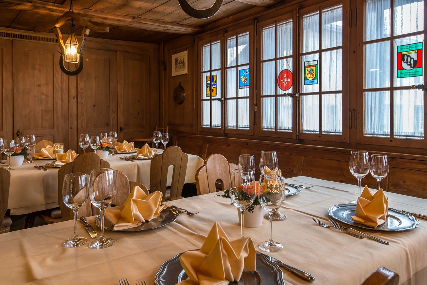 Ein festlich gedeckter Speiseraum mit mehreren Tischen, weißen Tischdecken, vorbereitet mit Gläsern, Besteck und gefaltenen gelben Servietten. Die Wände und Decke sind aus Holz, die Fenster sind groß mit durchsichtigen Vorhängen, und an den Fenster s