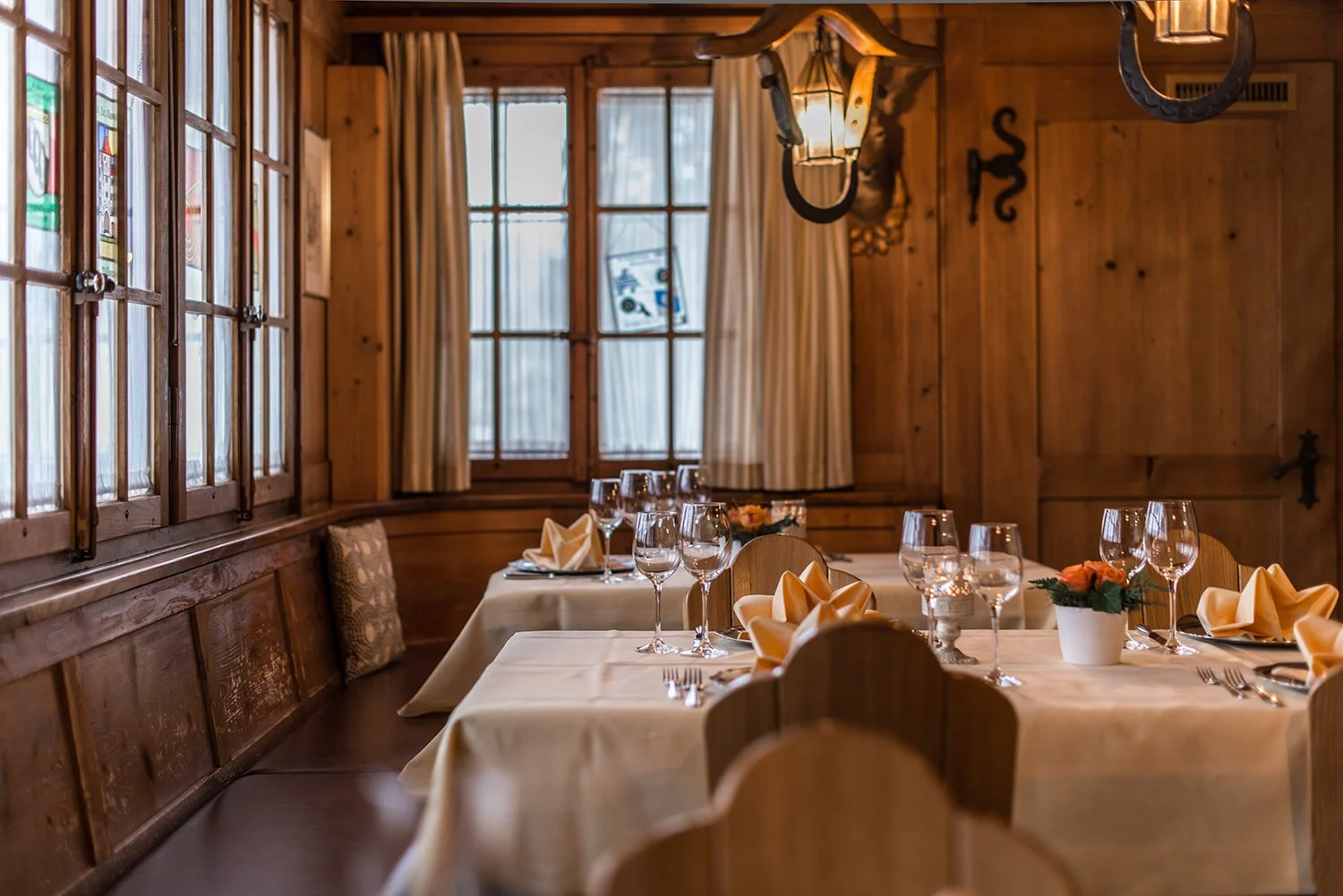 Ein rustikaler Speiseraum mit Holzvertäfelung, dekoriert für eine Mahlzeit mit Teller, Weingläser, Besteck, Stoffservietten, Kerzen und Blumenarrangements auf Tischen.