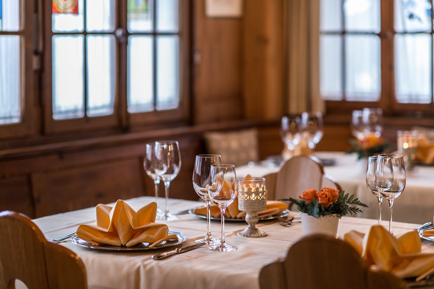 Ein festlich gedeckter Tisch in einem rustikalen Raum mit Holzvertäfelung, mit weißen Tischdecken, gefalteten gelben Servietten, Weingläsern, Kerzenlicht und einer kleinen Blumenarrangement.