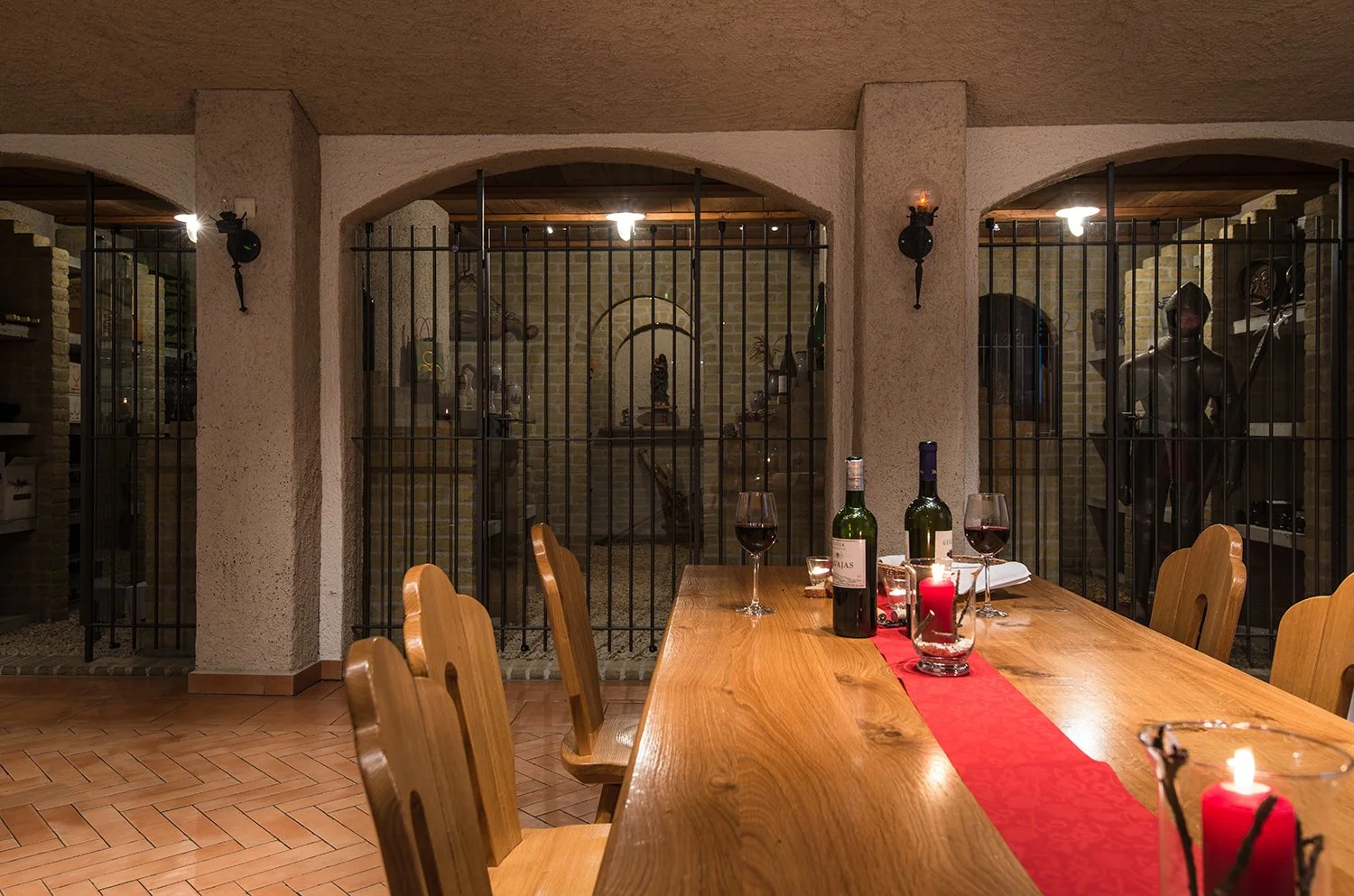Ein Esstisch mit Wein, Kerzen und einem roten Tischläufer in einem rustikalen Esszimmer, hinter einem Gittertor befinden sich Gegenstände und Skulpturen.