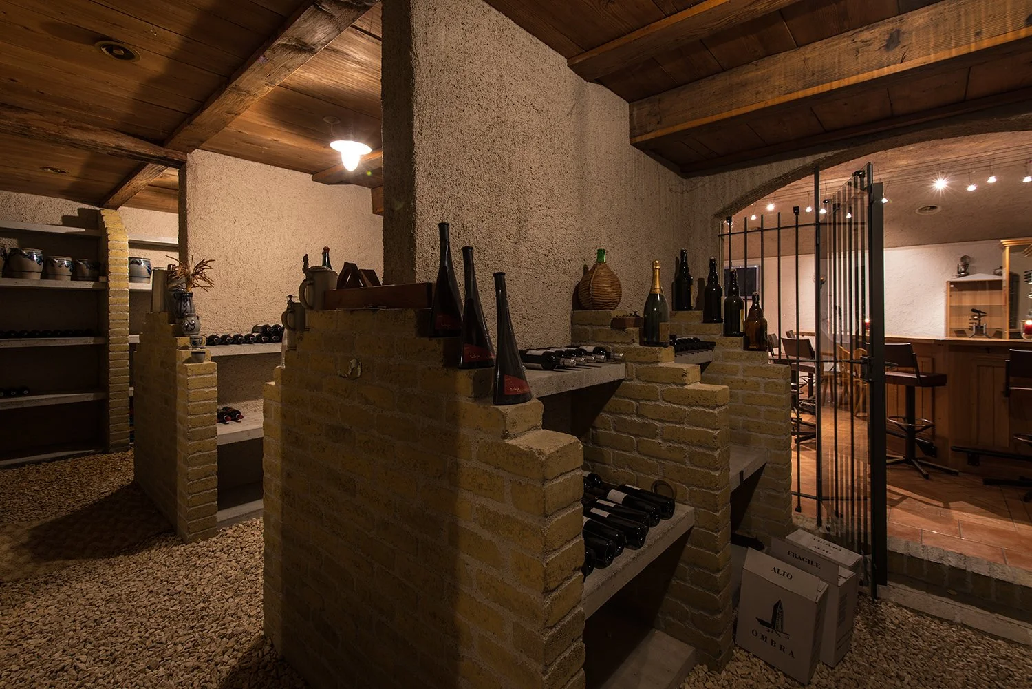 Ein gemütlicher Weinkeller mit Flaschen auf gestapelten Ziegelmauern, Holzregalen und einem angrenzenden Raum mit Bar und Barhockern. Der Raum ist warm beleuchtet, mit Holzboden und einer Holzdecke.
