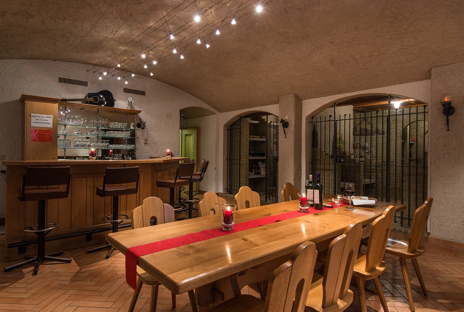Ein gemütlicher Speiseraum mit Holzmöbeln, einer langen Tafel, Kerzen und einem Barbereich im Hintergrund, beleuchtet mit warmem Licht.