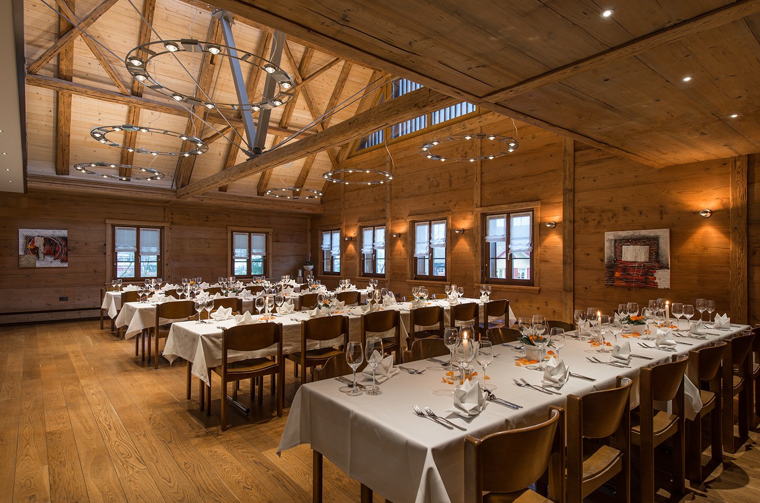 Ein festlich gedeckter Speisesaal in einem Holzhaus mit runden Kronleuchtern und großen Fenster.