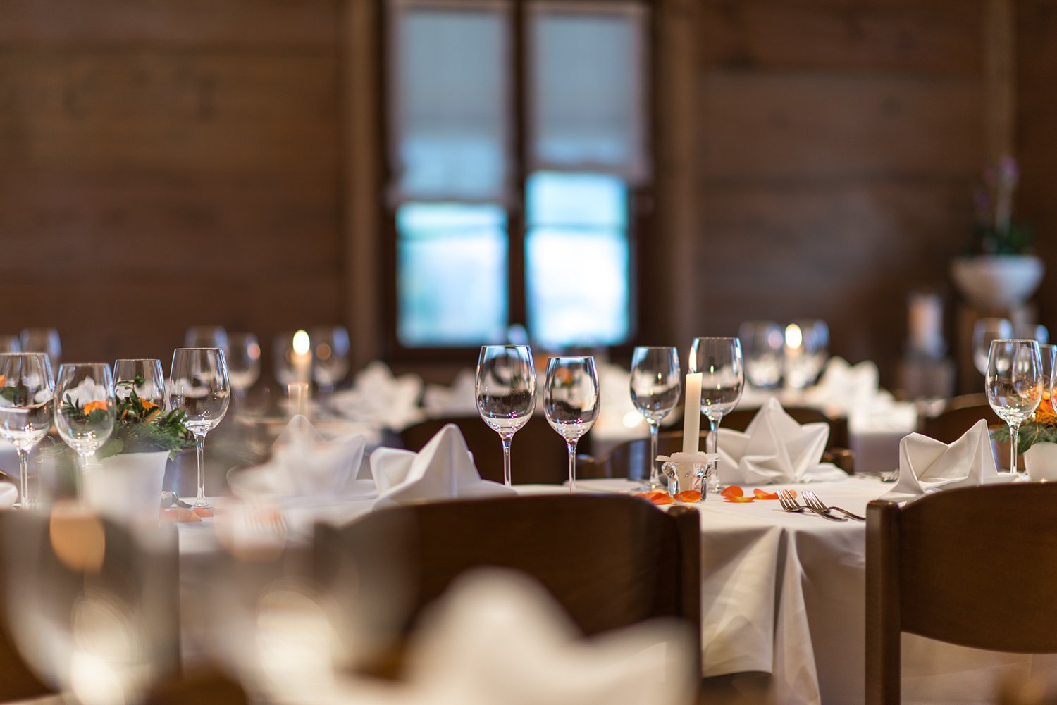 Ein festlich gedeckter Tisch in einem Raum mit Holzdekor, set mit Weingläsern, weißen Stoffservietten, Kerzen und Blumenarrangements.