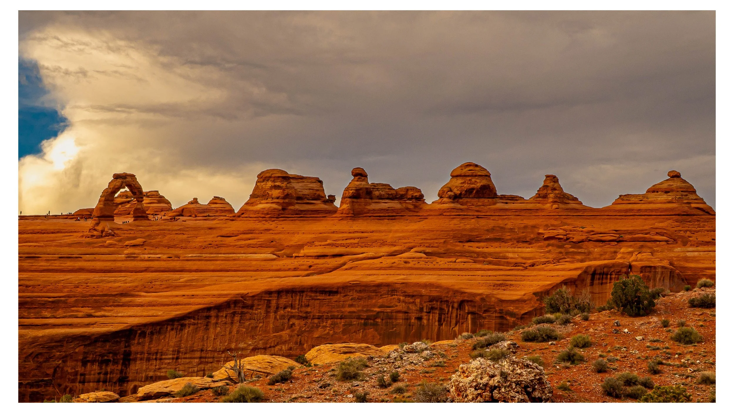 View of the desert landscape featuring large sandstone formations and the iconic rock arch with a cloudy sky overhead.