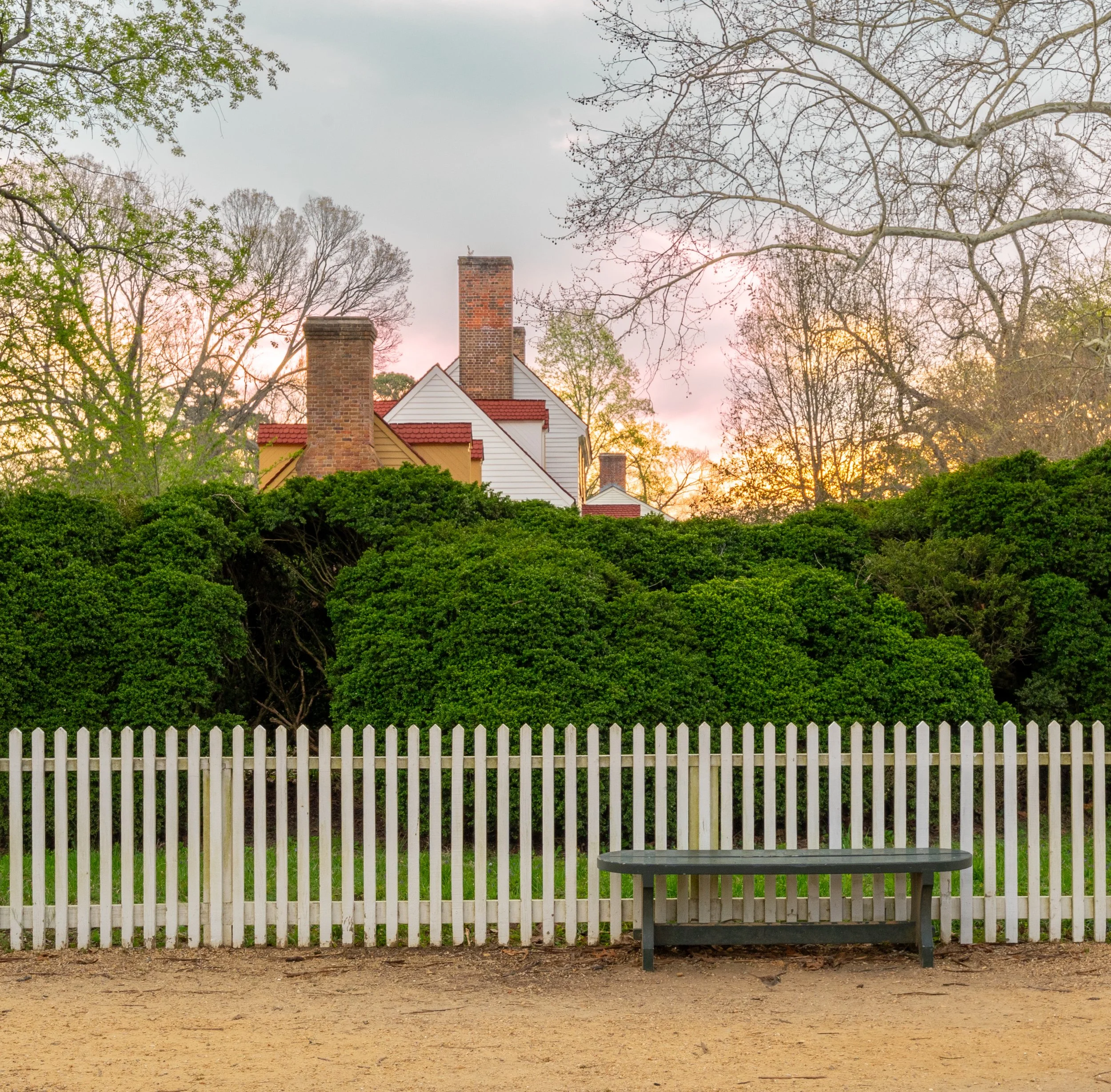 A white picket fence in the foreground with a gray park bench in front of it. Behind the fence, there are green bushes, leafless trees, and a house with a brick chimney and white siding. The sky is cloudy with some sunlight peeking through.