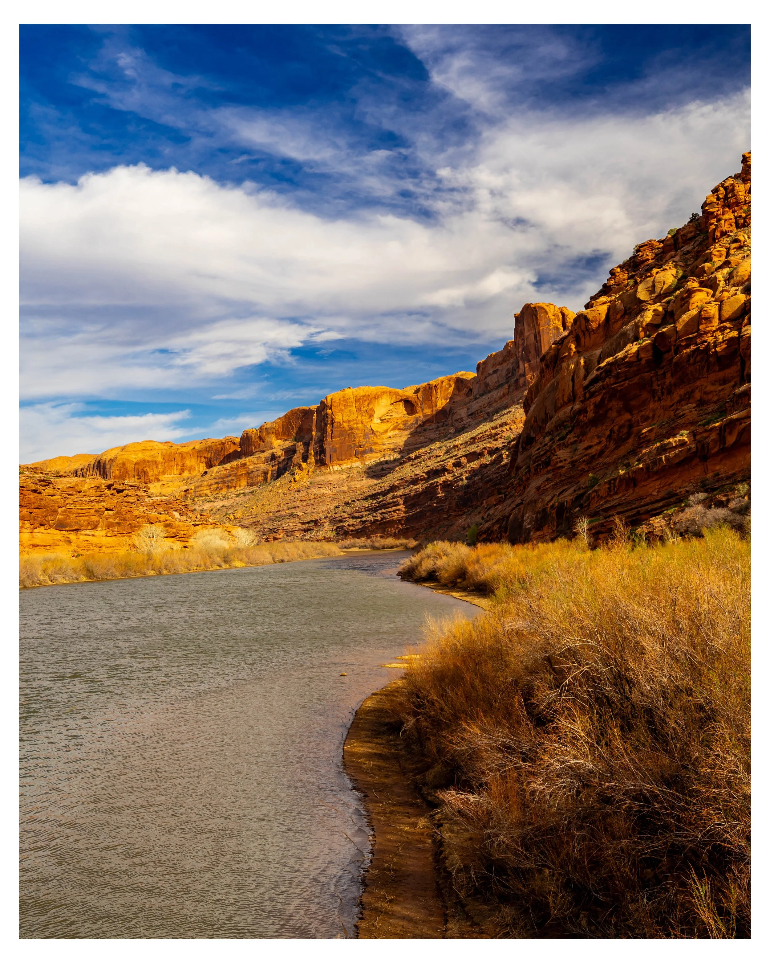 Scenic view of the Colorado River near Moab, Utah, winding through a desert landscape with rocky cliffs on the right and a blue sky with clouds above.