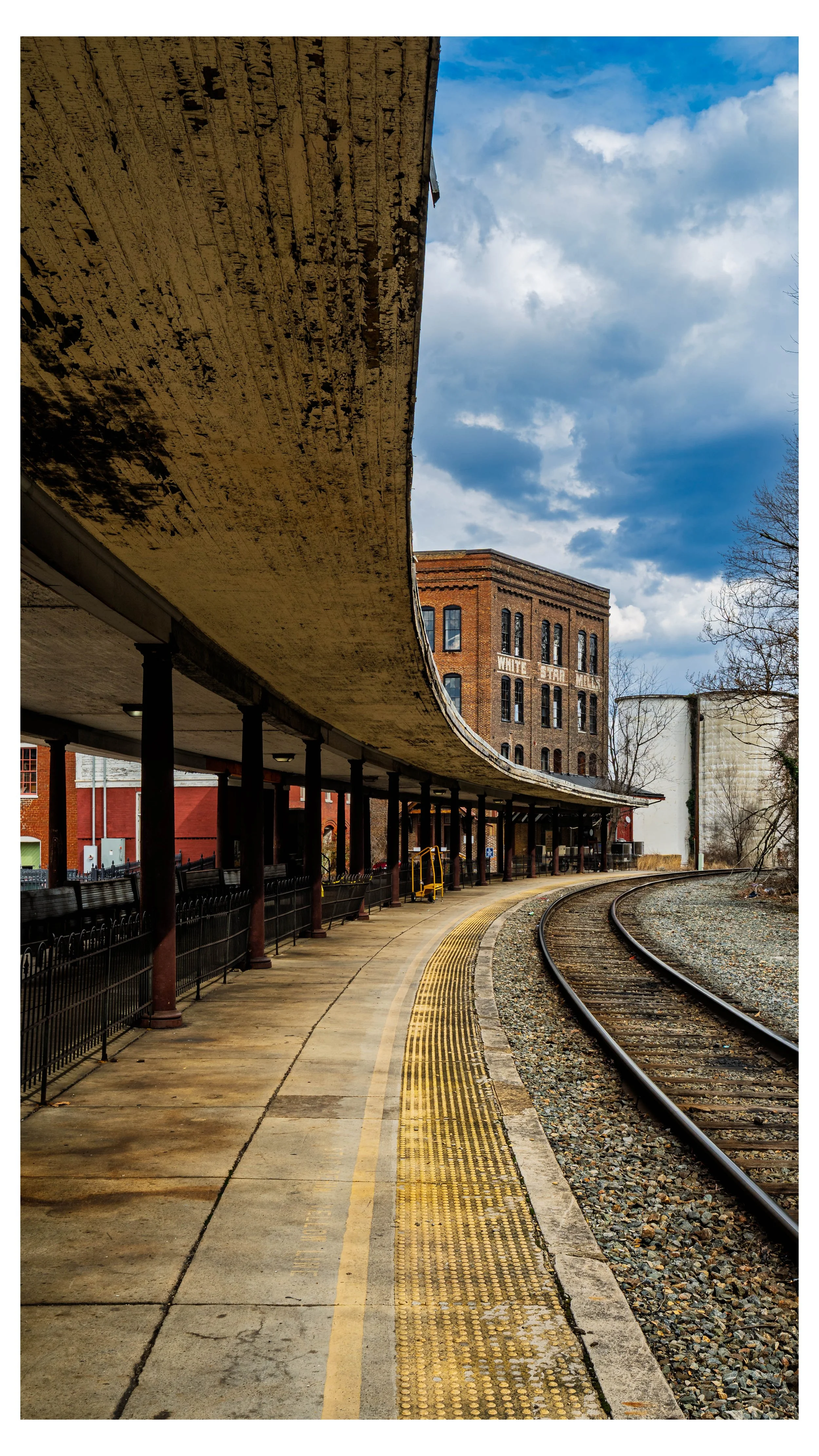 Staunton, Virginia's curving train platform with roof, railroad tracks, and historic brick building in the background under a cloudy sky.