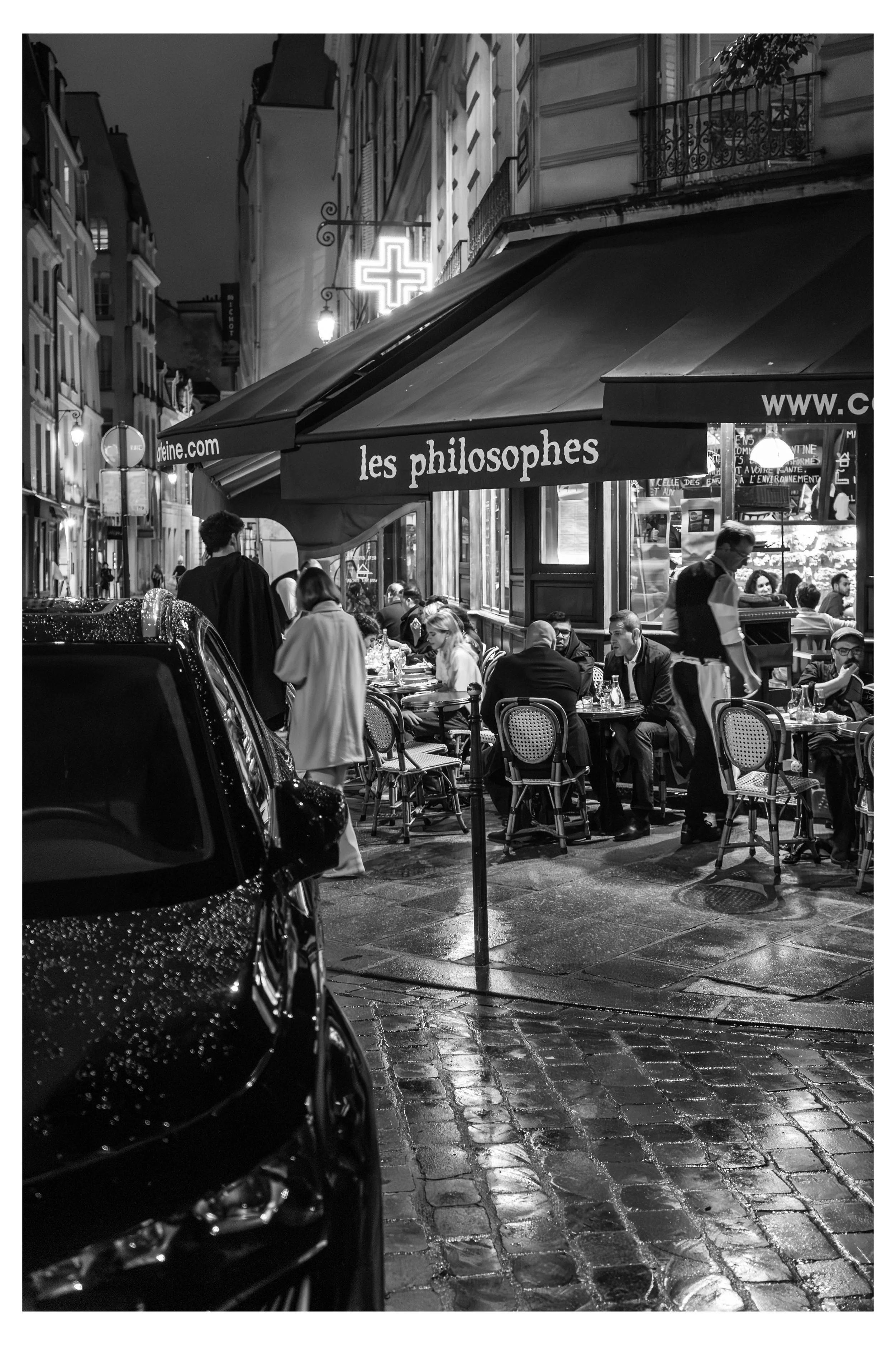 Nighttime scene at a Paris street café with outdoor seating, people dining, a neon cross sign indicating a pharmacy, and buildings with classic architecture.