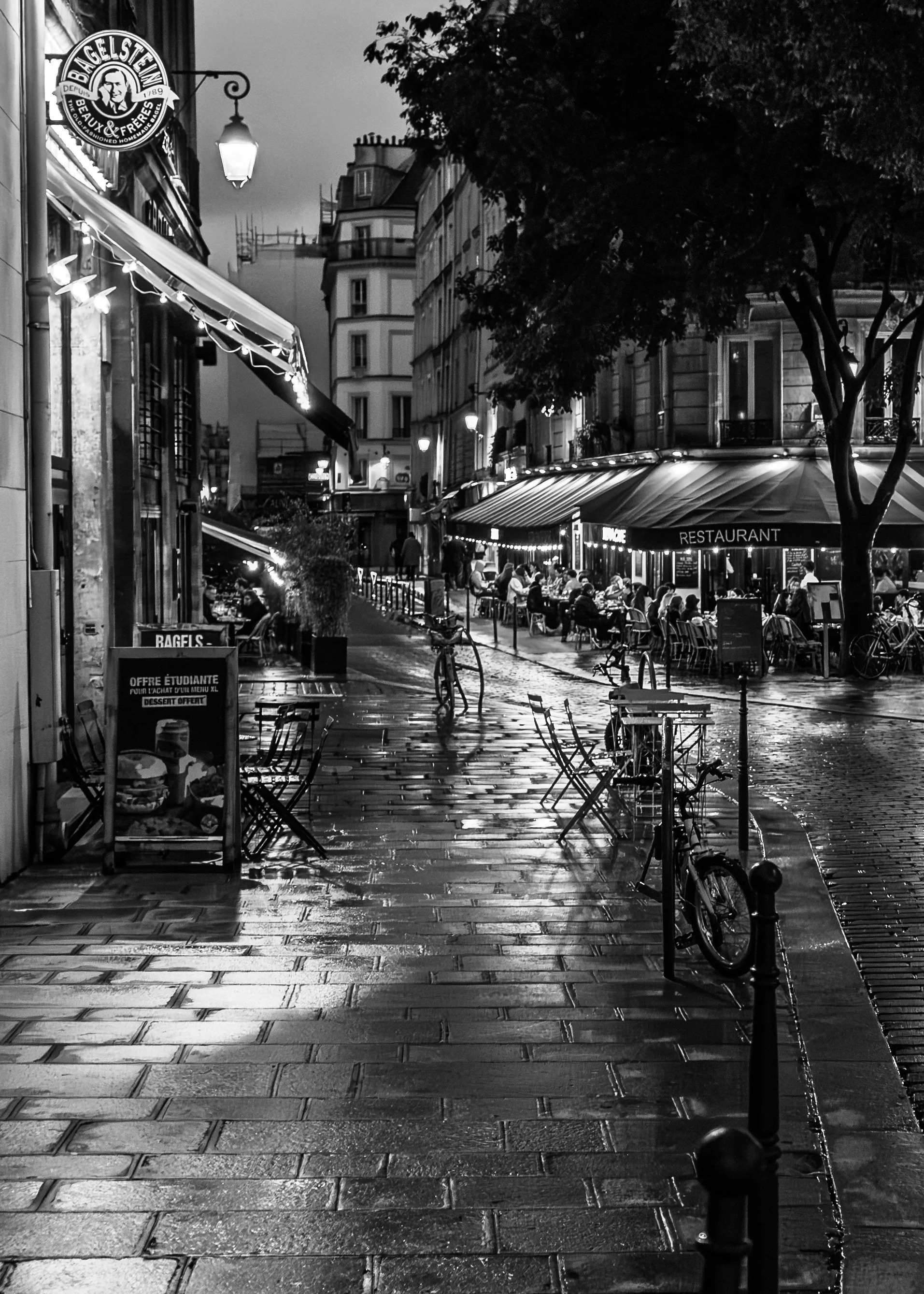 A nighttime street scene with outdoor dining, bicycles, and street lamps, in Le Marais District, Paris. 