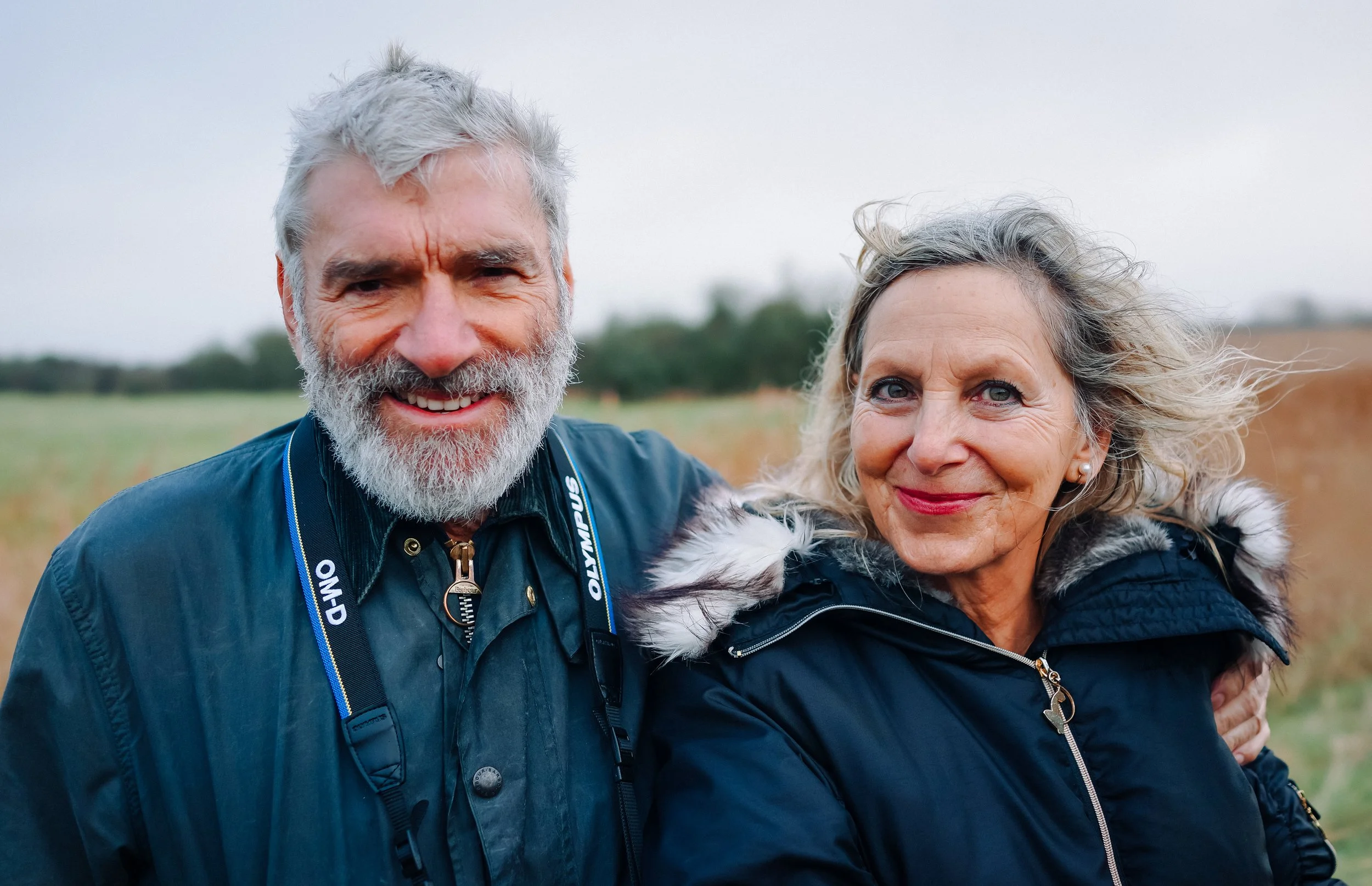 A smiling elderly man with gray hair and beard and an elderly woman with gray hair, both wearing warm jackets, standing outdoors in a field with a cloudy sky in the background.