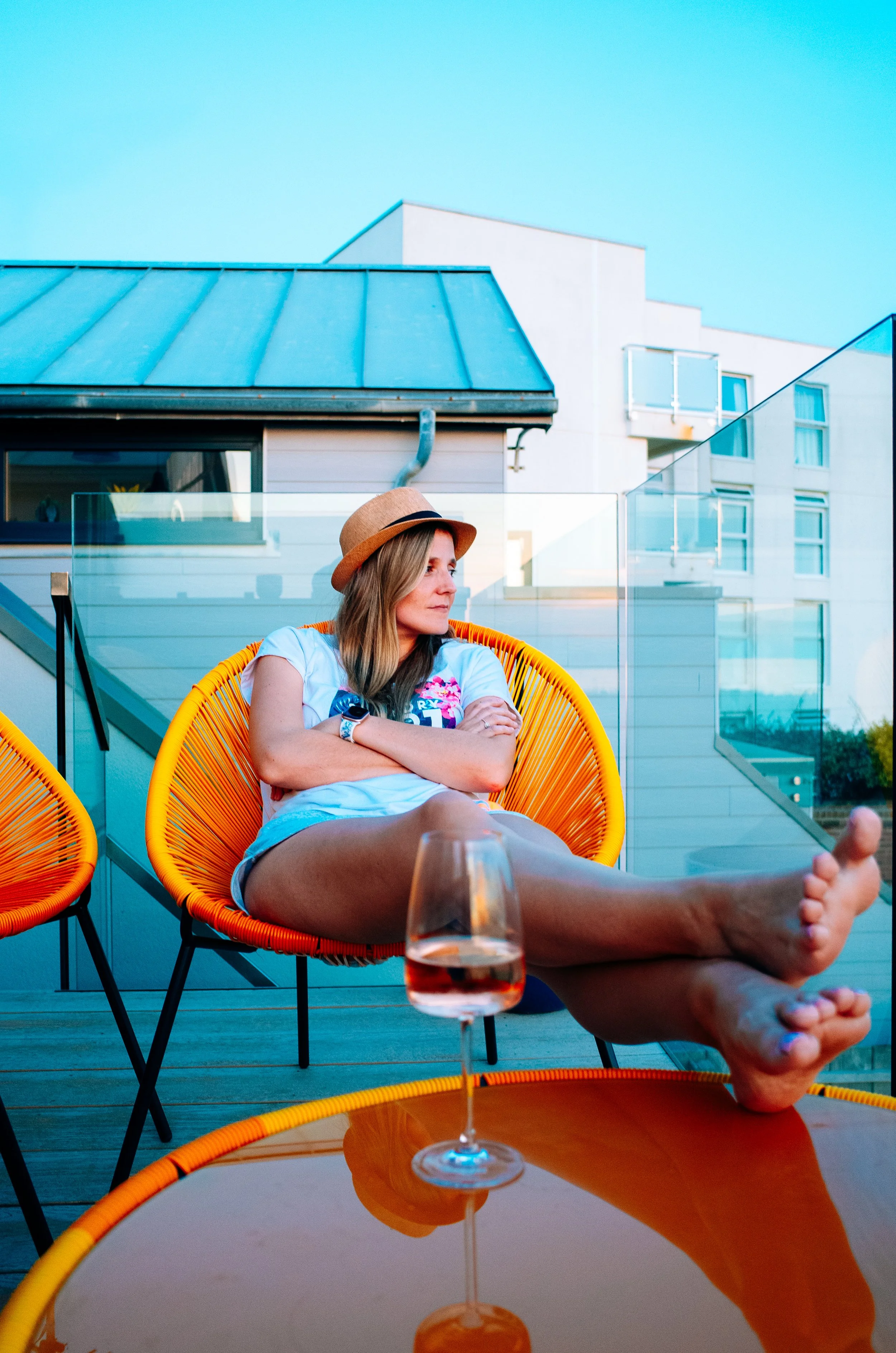 A woman wearing a tan hat and white t-shirt, sitting outdoors on a yellow modern chair, with a glass of rosé wine on a reflective table in front of her. She has arms crossed and looks thoughtfully to her left. The background features balconies and ap