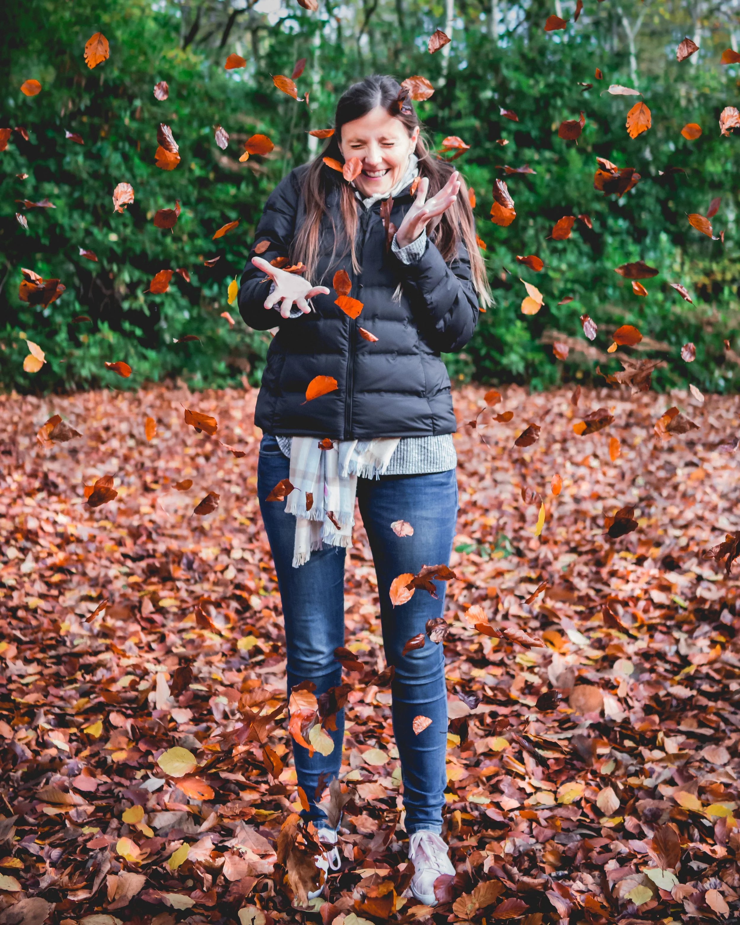 A woman with long dark hair, wearing a black jacket, grey sweater, and blue jeans, is standing in an autumn forest with fallen leaves on the ground. She is laughing and joyfully tossing orange and brown leaves into the air.