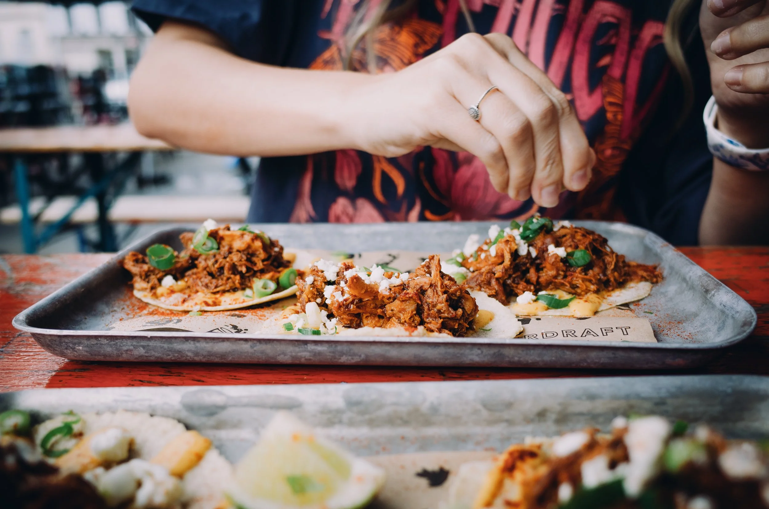 Close-up of hands with a ring, preparing or serving tacos with shredded meat, topped with chopped green onions, and cheese on a tray, on a wooden table in a restaurant.