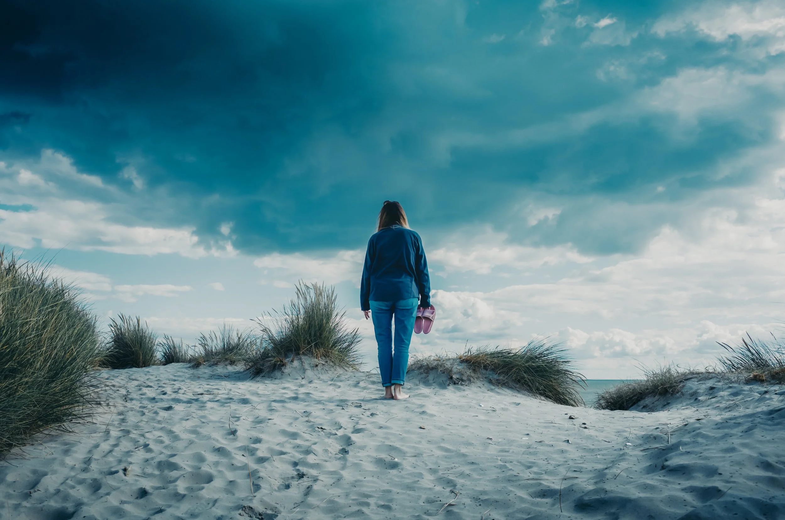Woman walking on beach holding pink shoes, with cloudy sky overhead and sand dunes with grass nearby.