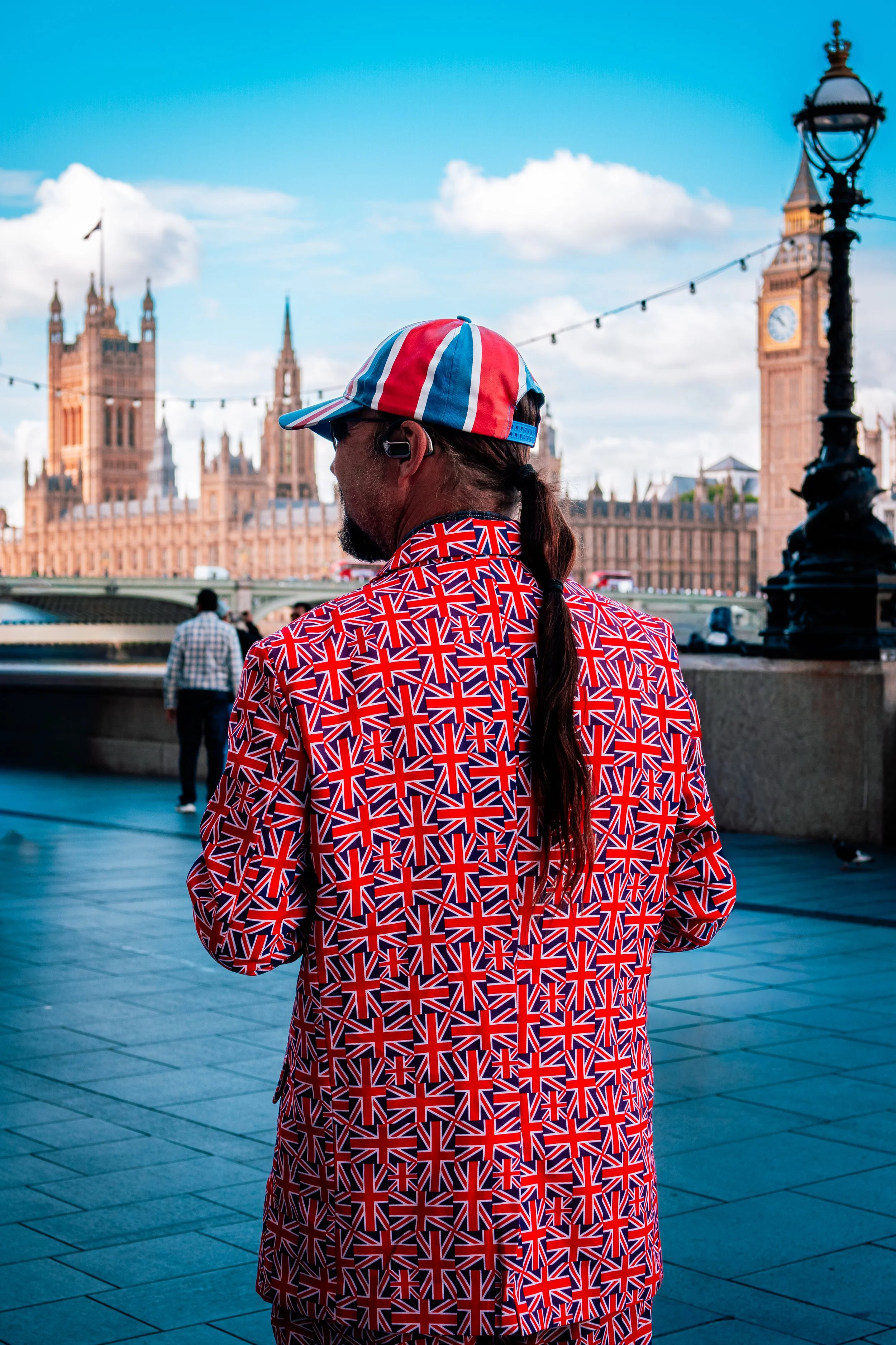 Person with long hair wearing a Union Jack themed jacket, cap, and Bluetooth earpiece, standing outdoors in front of the Houses of Parliament and Big Ben in London, UK, on a sunny day.