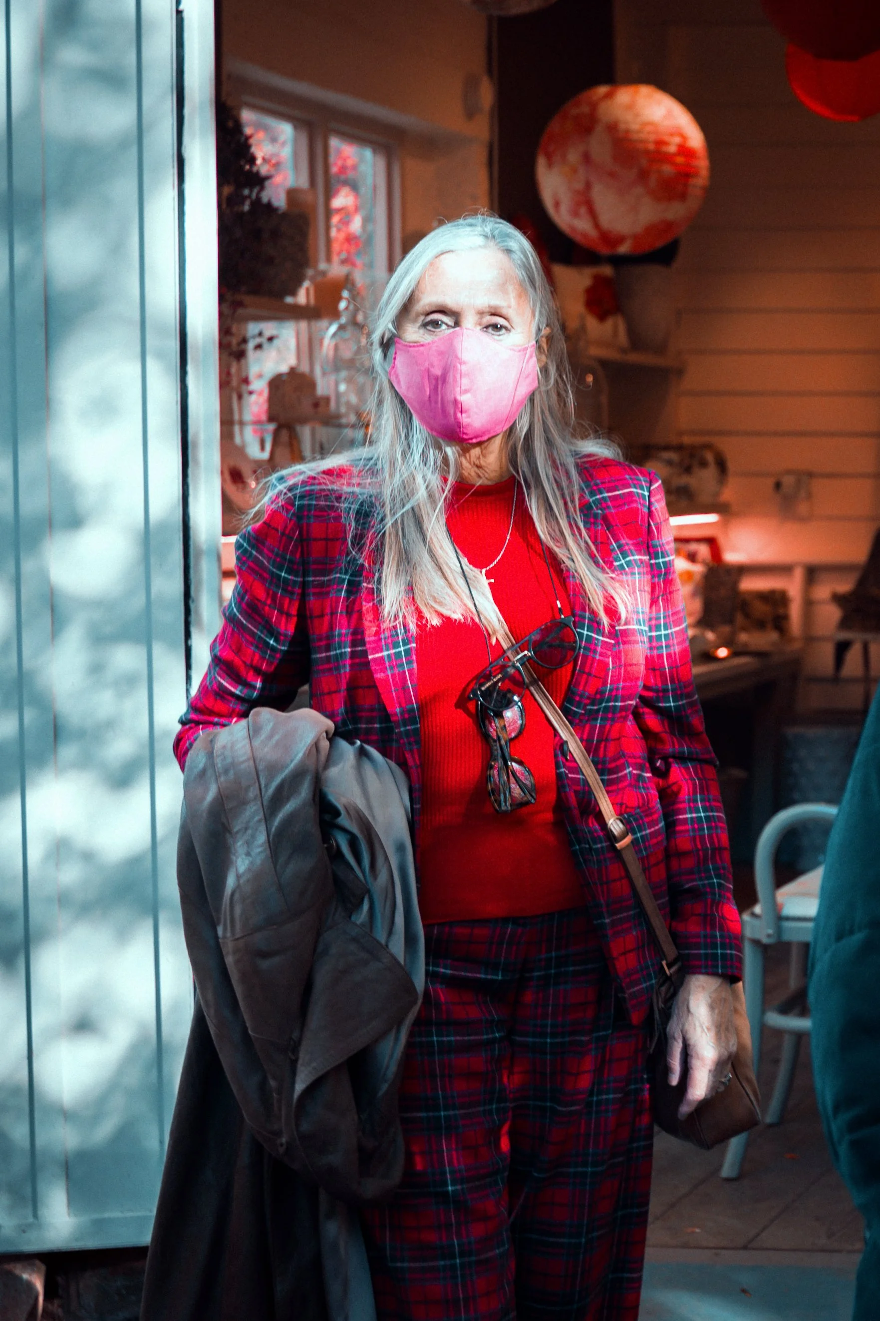 An elderly woman wearing a pink face mask, red sweater, and red plaid blazer standing indoors near a window, with decorative paper lanterns hanging overhead.