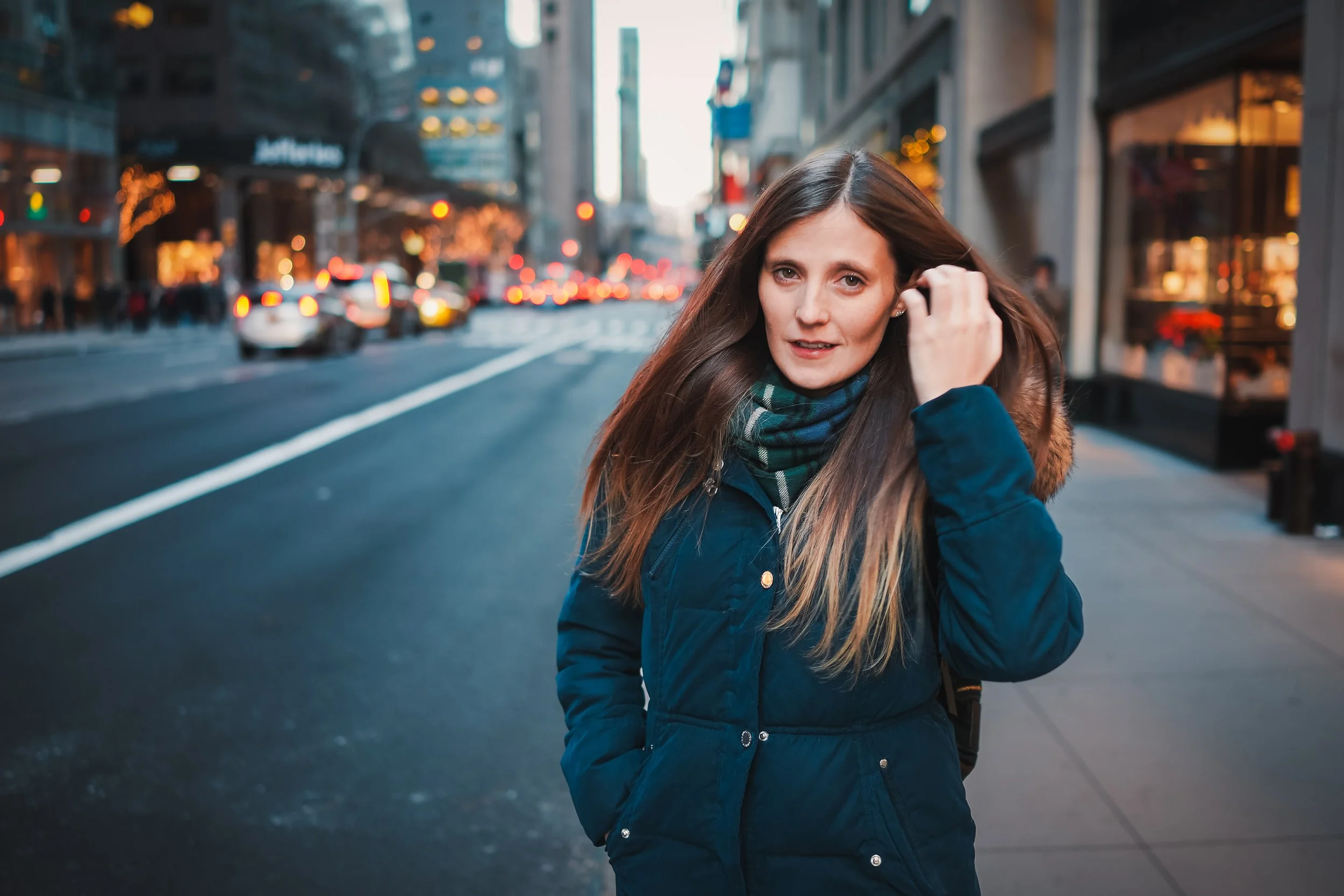A woman with long brown hair, wearing a navy blue coat and a plaid scarf, is standing on a city street during dusk, adjusting her hair. The background shows blurred city lights, buildings, and cars.