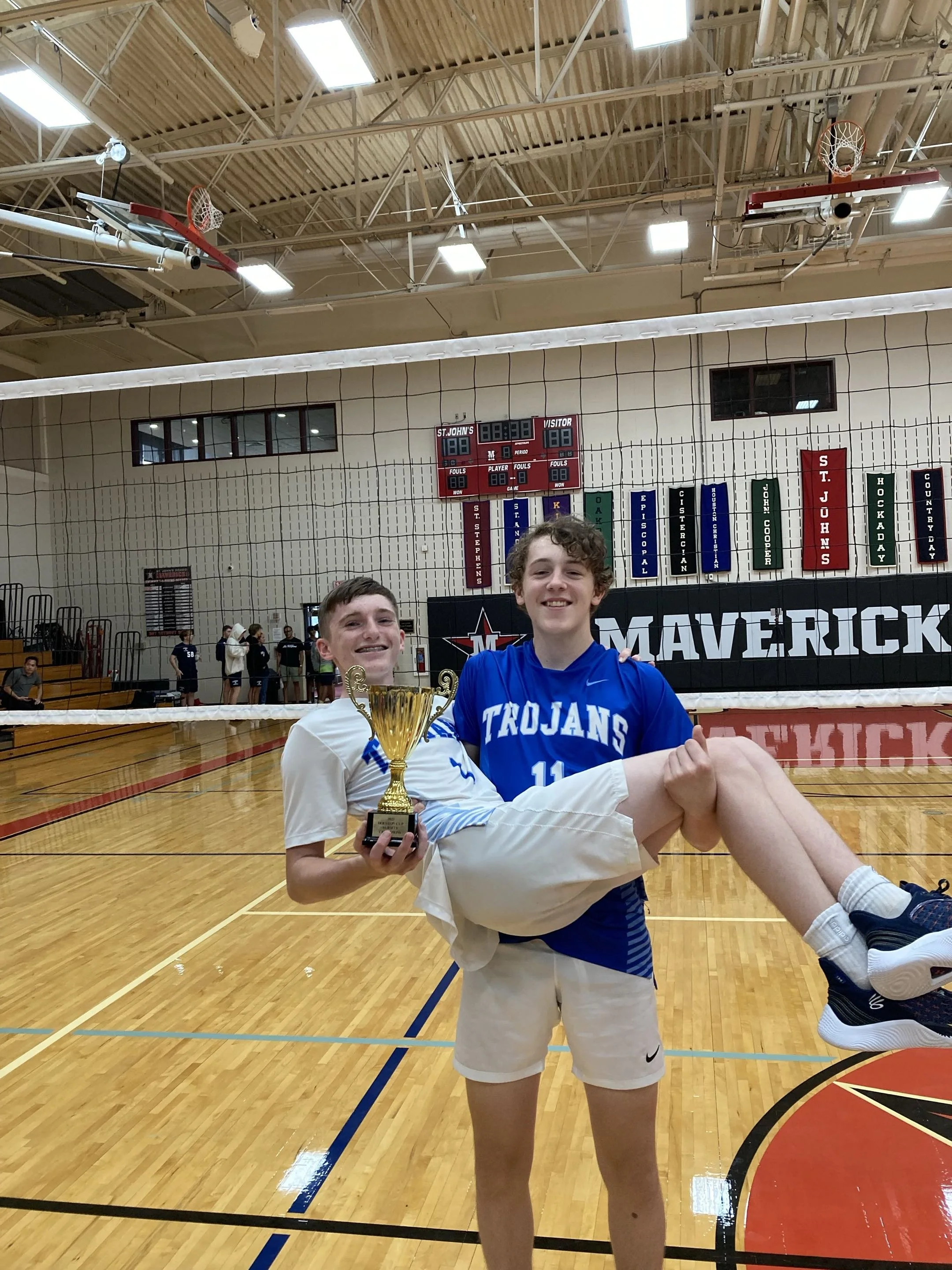 Two young boys in a volleyball gym, holding a trophy. One boy is wearing a white and blue volleyball uniform, and the other is in a blue jersey with 'Trojans' and the number 11. They are smiling, celebrating a win, with a volleyball net, a scoreboard, and banners in the background.