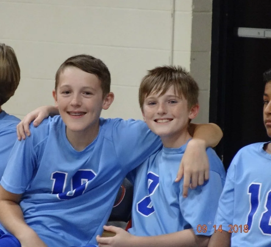 Two young boys in blue soccer jerseys sitting close together, smiling, with their arms around each other, in a team setting.