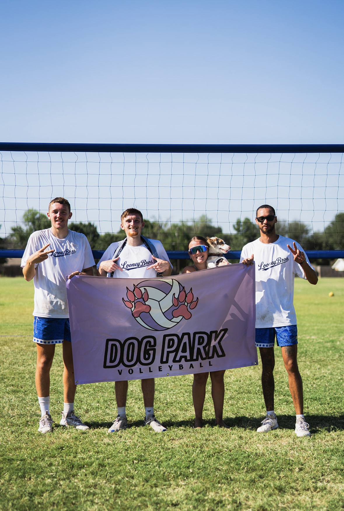 Group of four people and a dog holding a volleyball-themed banner that reads 'Dog Park Volleyball' on a grassy field with a volleyball net behind them.
