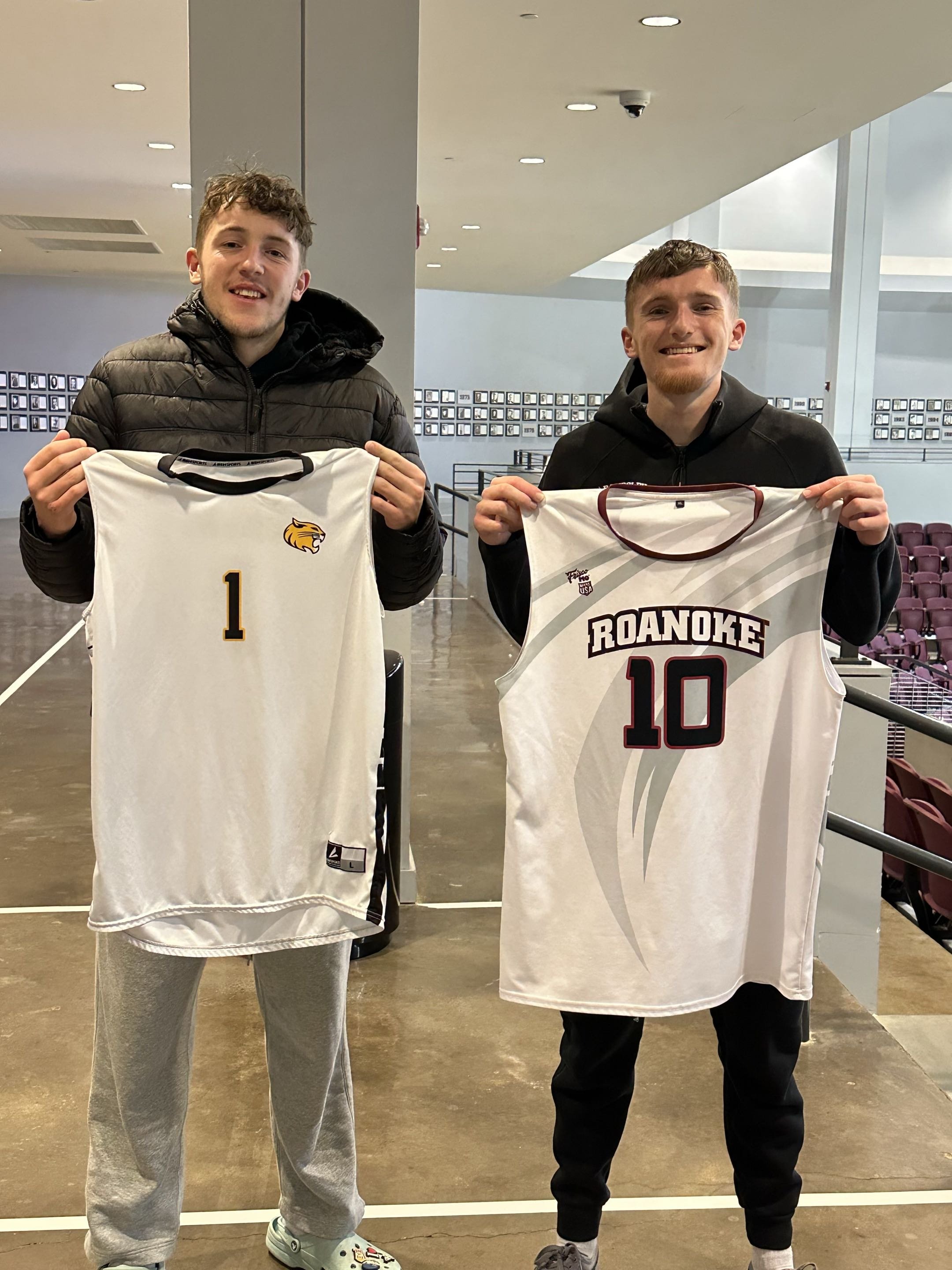 Two young men smiling and holding basketball jerseys indoors, with one displaying a jersey with number 1 and tiger logo, and the other with number 10 and Roanoke written on it.