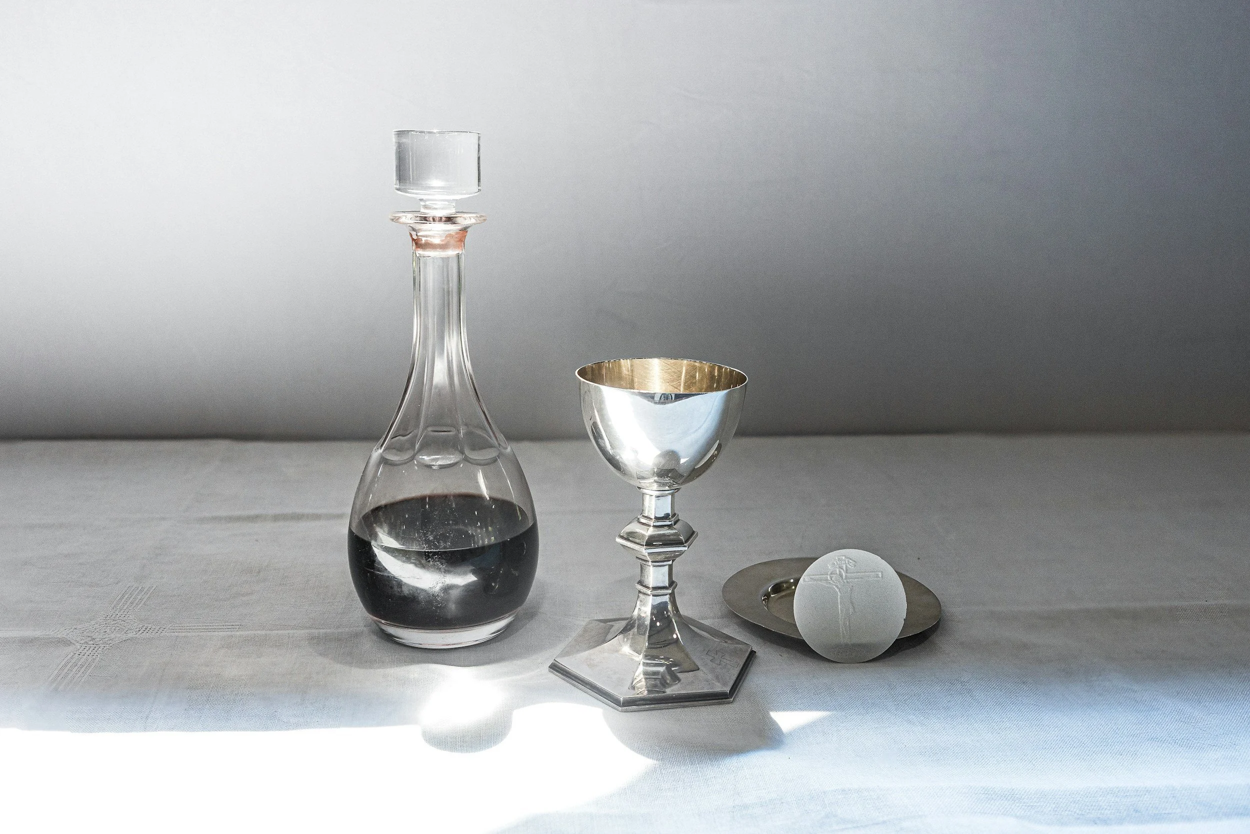 A glass decanter partially filled with dark liquid, a small silver bowl, and various glassware or metallic objects on a white tablecloth with a gray wall background.