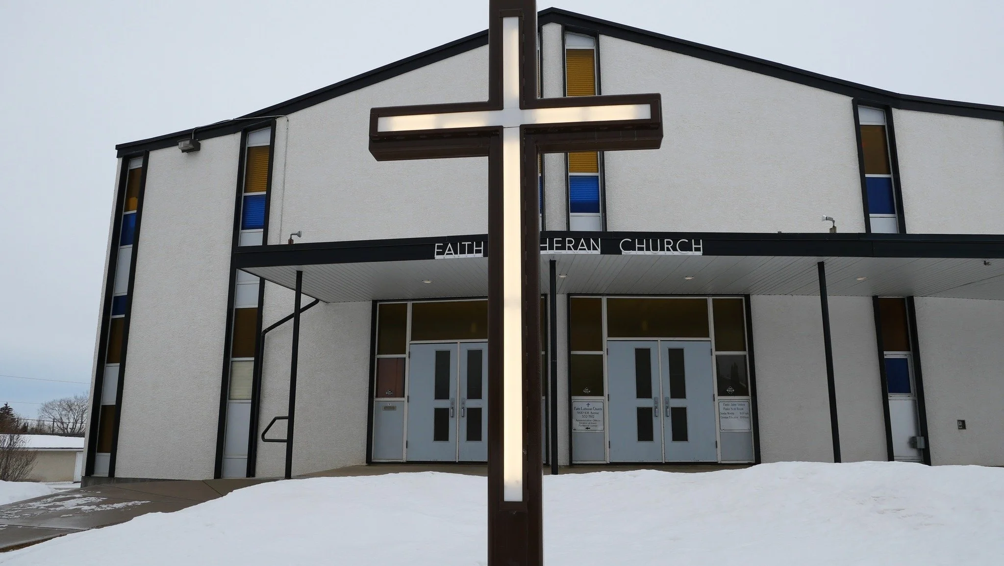 Exterior of a modern church building called Faith Lutheran Church with stained glass windows and a cross sign in front, surrounded by snow.