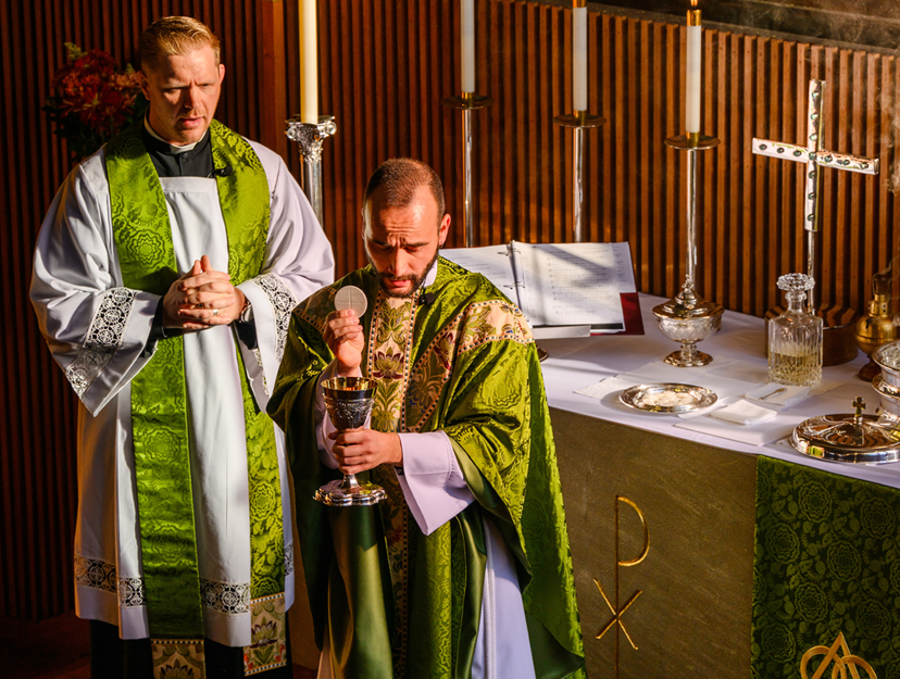 Two priests in green vestments at altar, one holding a chalice and wafer, the other praying, with lit candles and religious items on altar in a church.