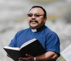 A smiling man dressed as a priest holding an open book.