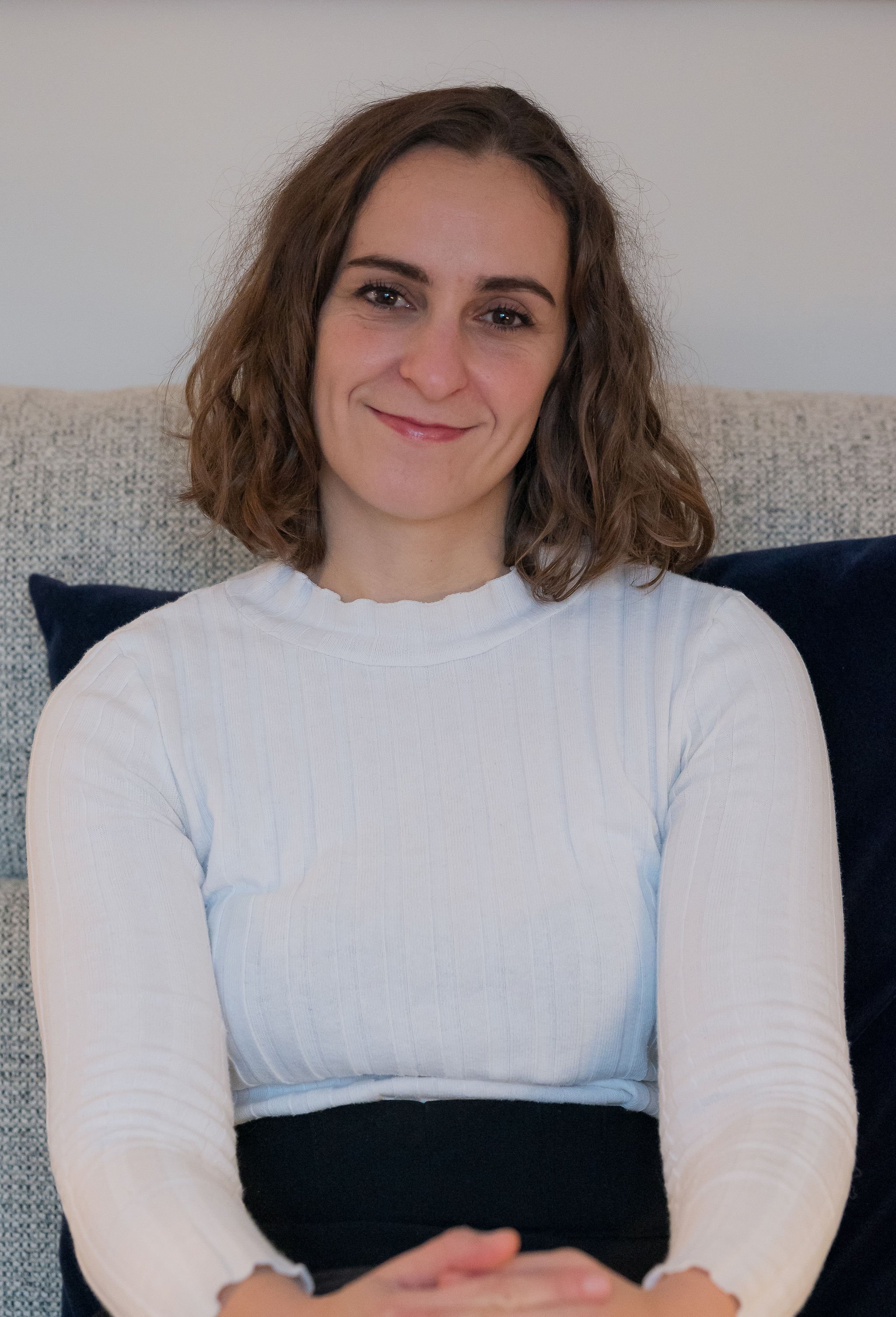 Woman with brown hair smiling, sitting on a couch with a dark pillow behind her, wearing a white ribbed long-sleeve top.