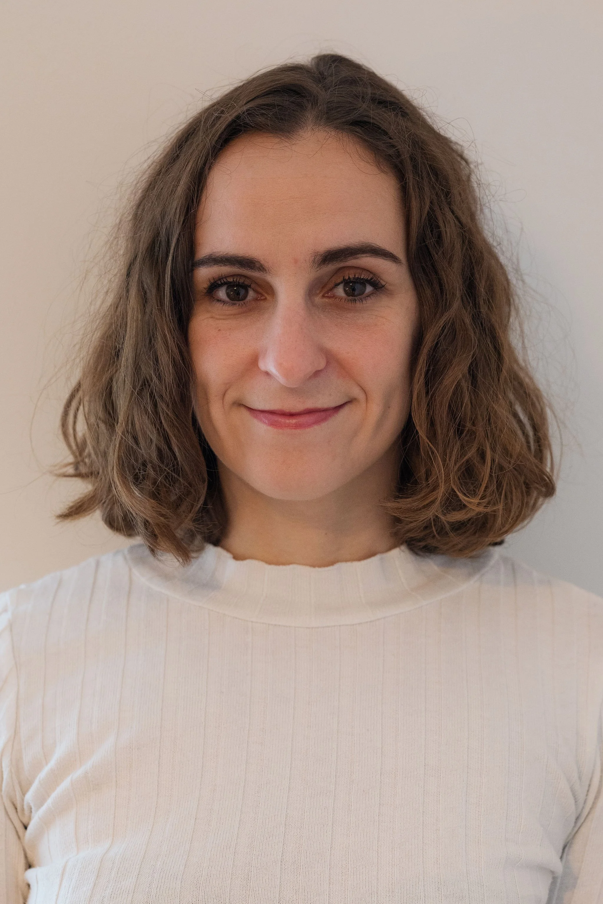 Close-up of a woman with curly brown hair, fair skin, and a subtle smile, wearing a white ribbed sweater, standing against a plain light-colored wall.