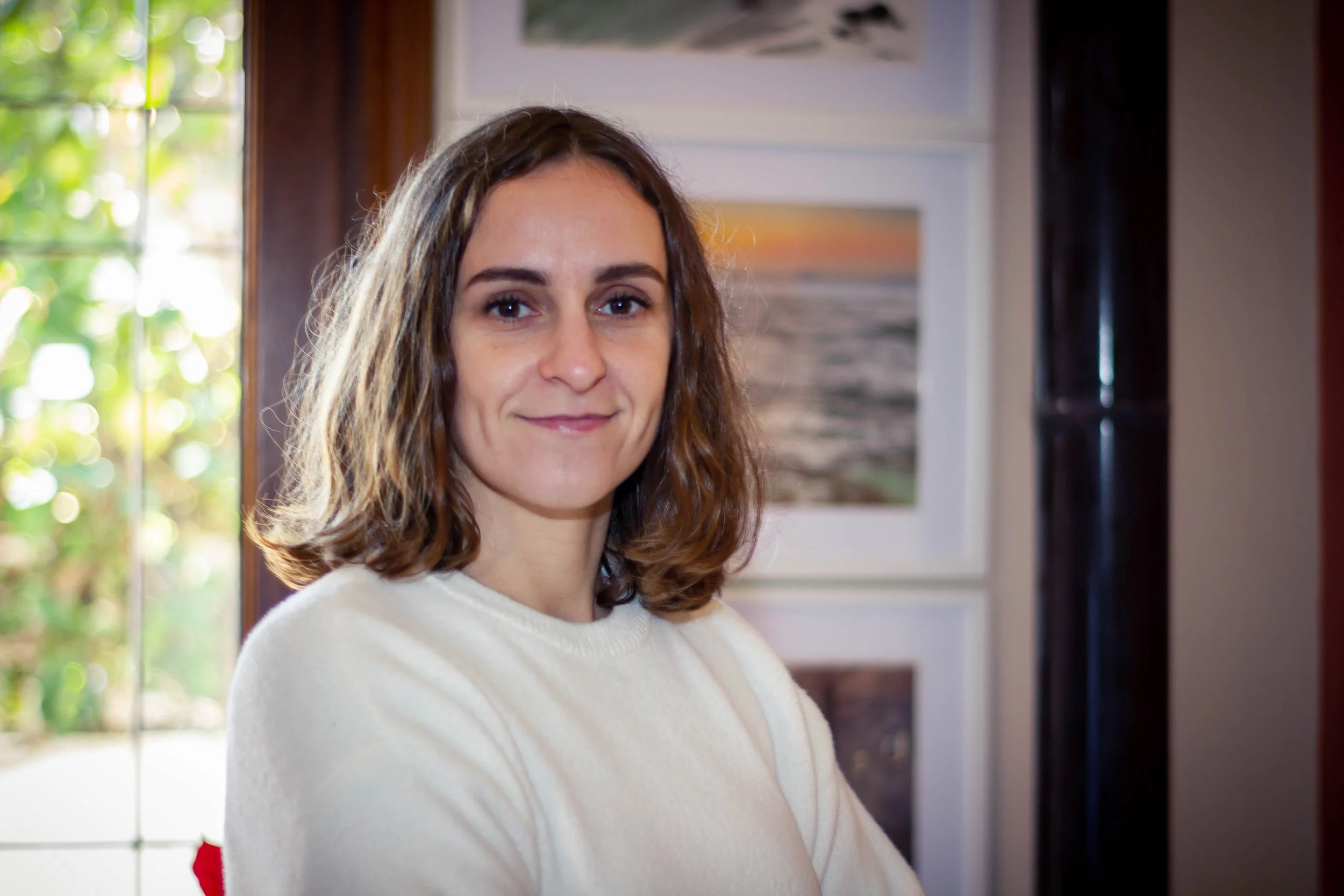 A woman with shoulder-length brown hair wearing a white sweater, standing indoors near a window with greenery outside, and artwork on the wall behind her.