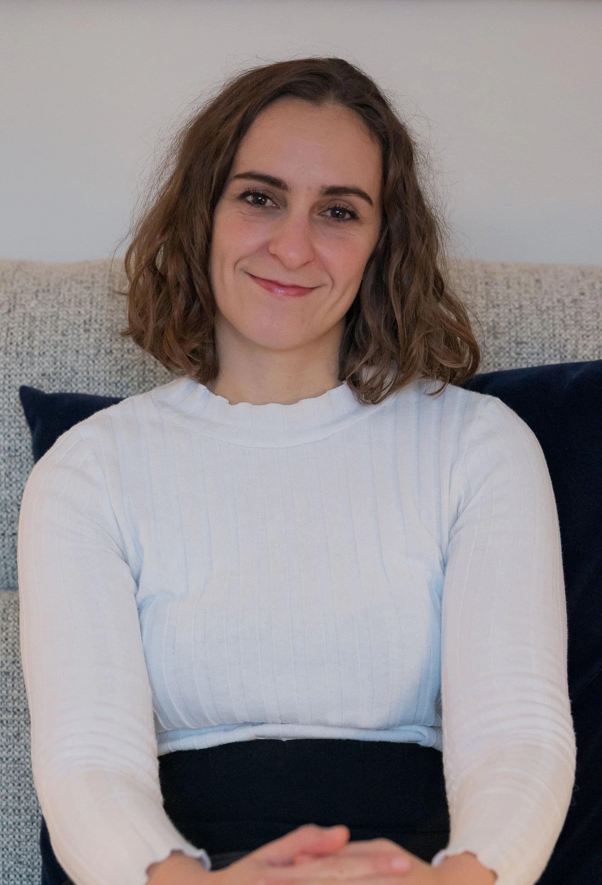A woman with shoulder-length brown hair, smiling, wearing a white long-sleeve ribbed shirt and sitting in front of a neutral wall with a beige couch and dark pillow behind her.