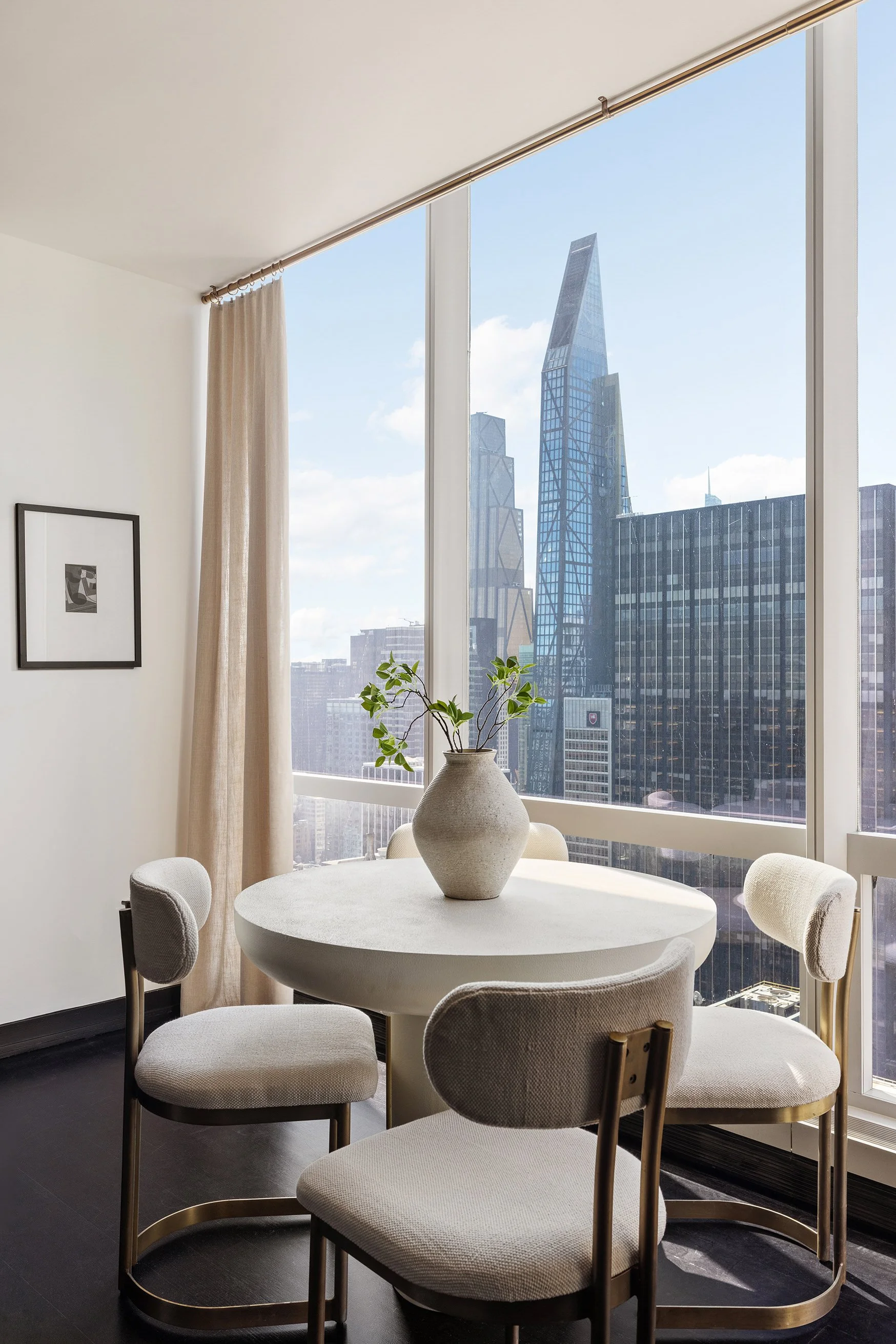 Modern dining area with a round table and six beige chairs, a large window with a city skyline view, a potted plant, and a framed abstract art on the wall.