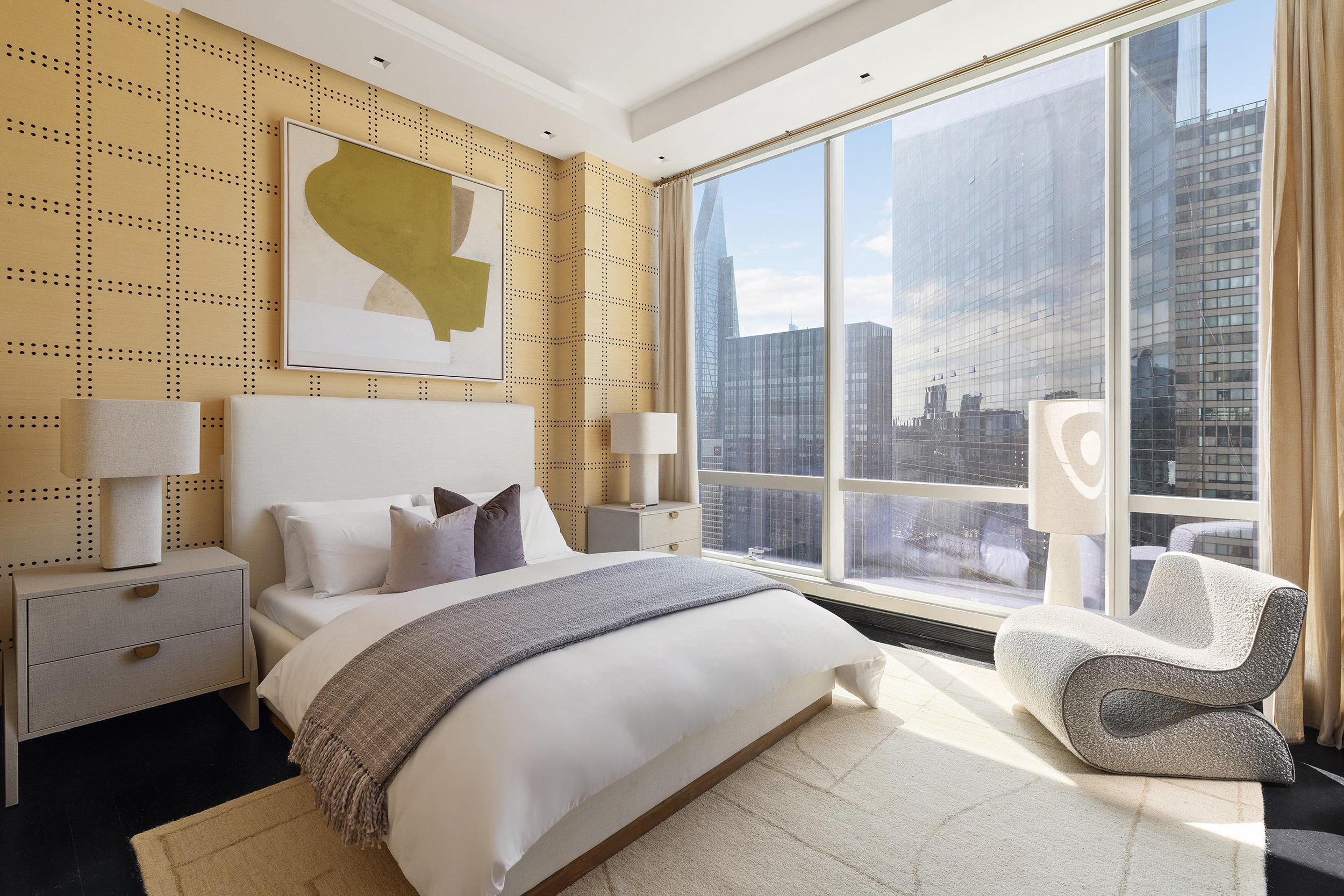 Modern bedroom with large window showing cityscape. White bed with gray throw, beige nightstand with lamps, abstract artwork on a dotted yellow wall, and a white upholstered chair.