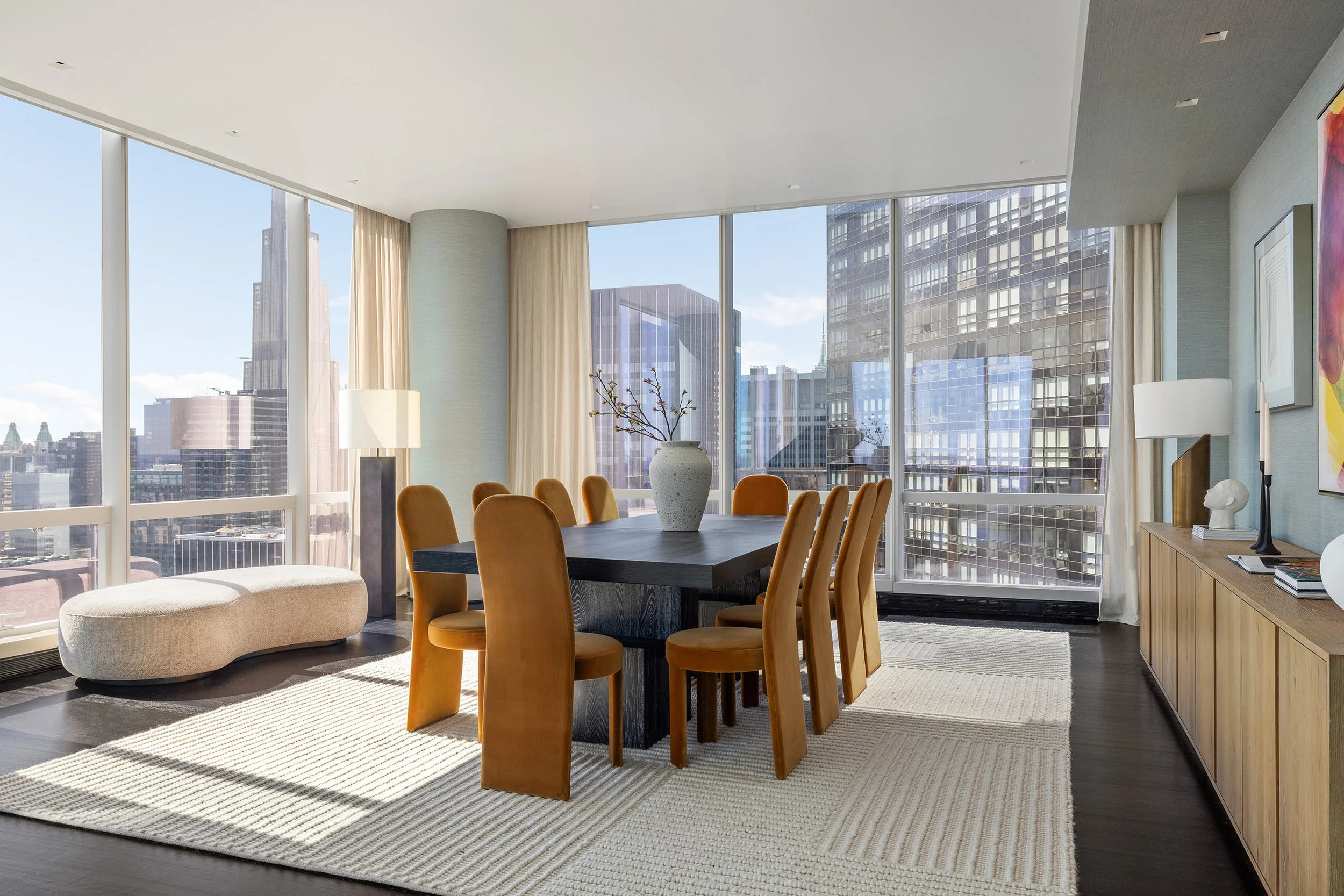 Modern high-rise apartment living room with large floor-to-ceiling windows offering city views, a dark wooden dining table with tan upholstered chairs, a white oval ottoman, a beige area rug, a tall white table lamp, a ceramic vase with branches, and