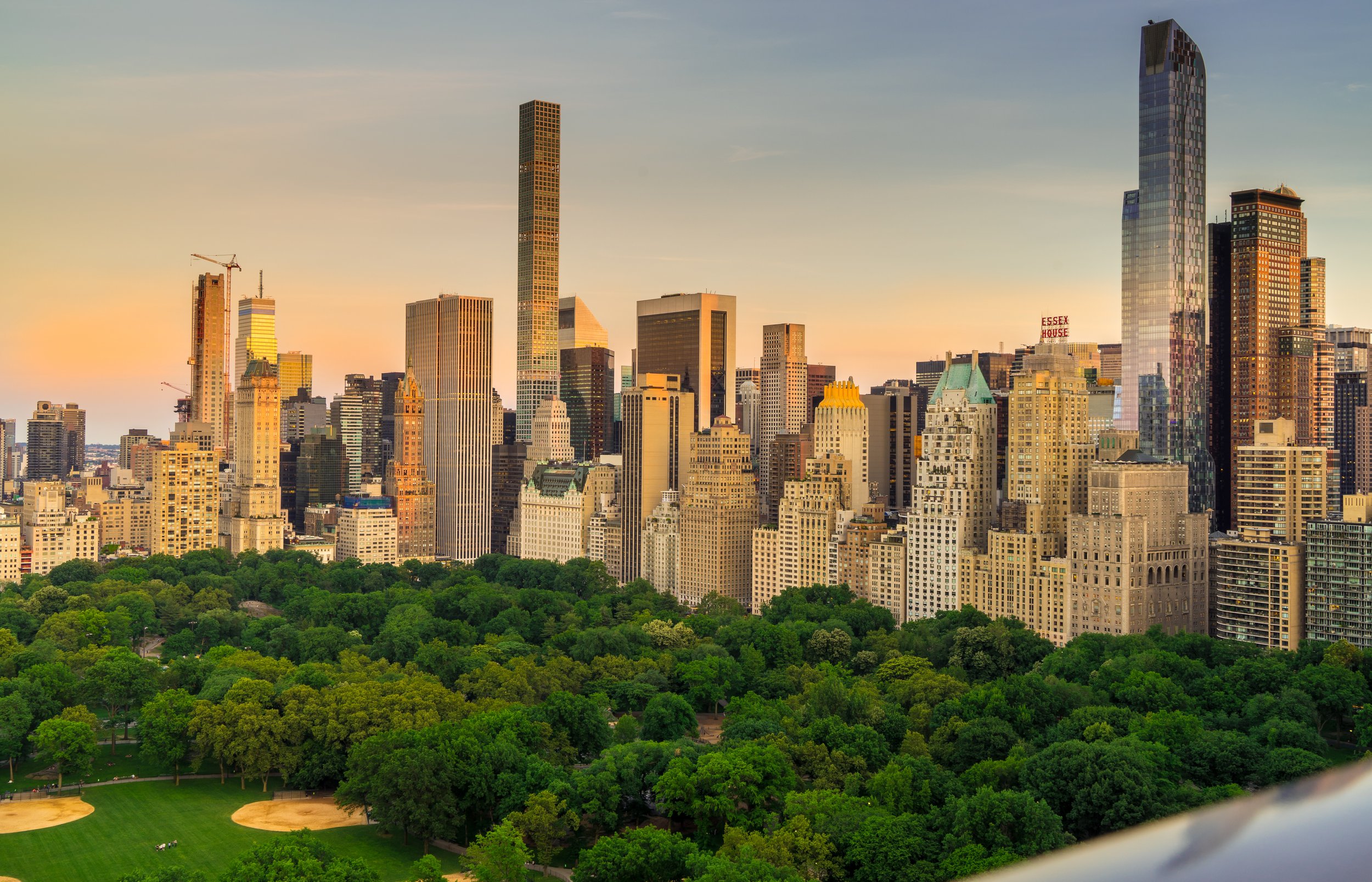 A city skyline at sunset with tall skyscrapers above a park with lush green trees, a baseball field, and winding pathways.
