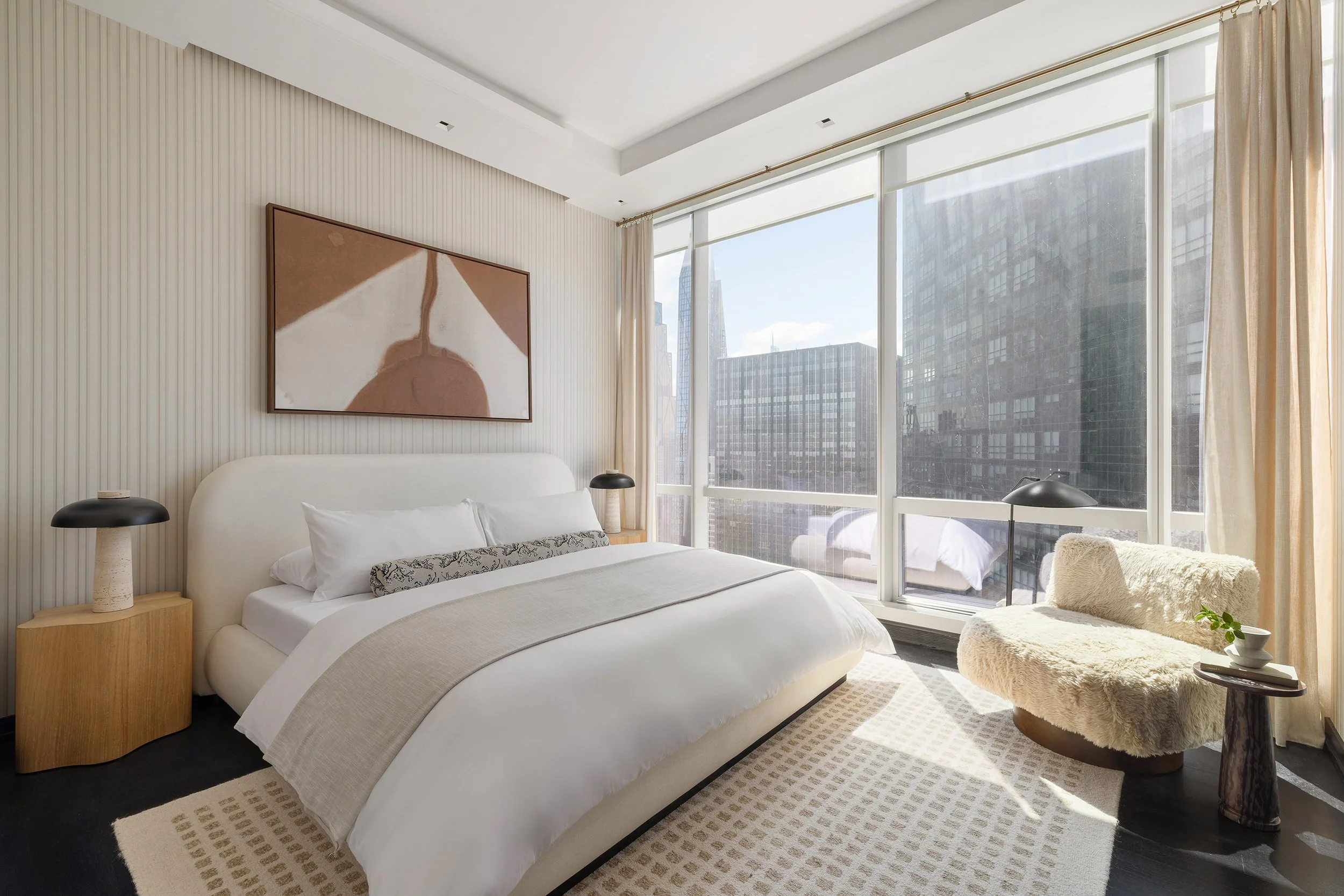 Modern bedroom with large windows, white bed, abstract wall art, and a cozy fur chair with a small table and plant.