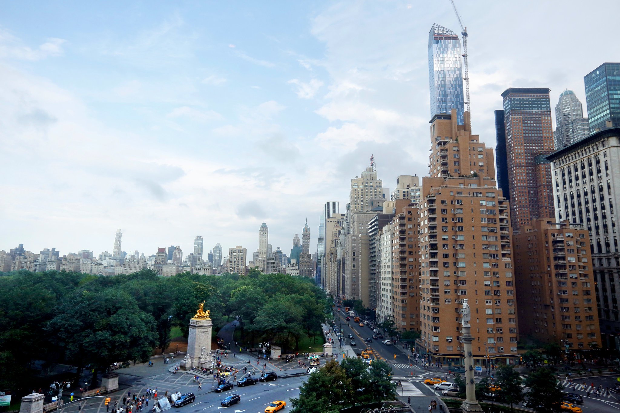 A cityscape with tall skyscrapers, a green park area with trees in the foreground, and a busy street with cars and pedestrians.