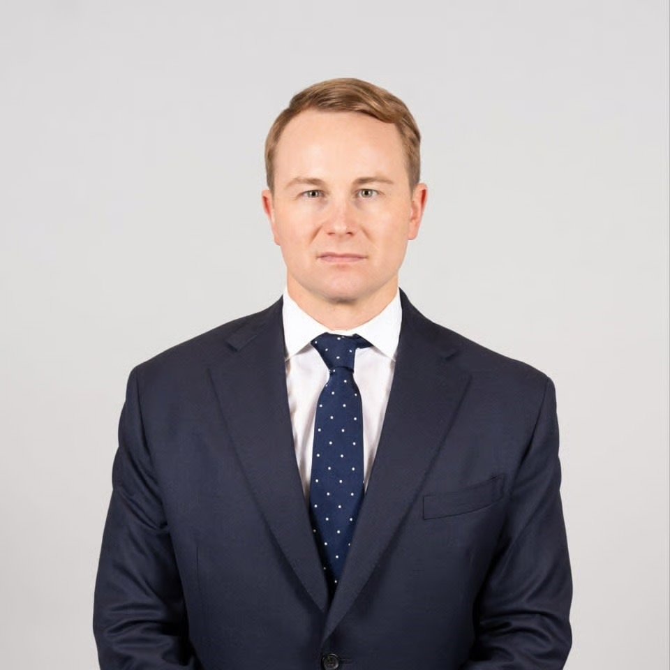 A man in a navy suit, white shirt, and navy tie with white polka dots standing against a plain light gray background.