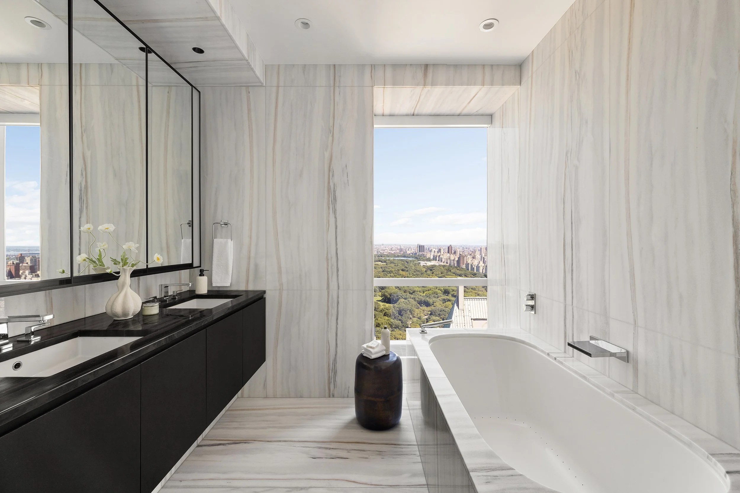Modern bathroom with white marble walls and floor, large window with city and park view, black vanity with two sinks, mirror, vase with flowers, and bathtub.