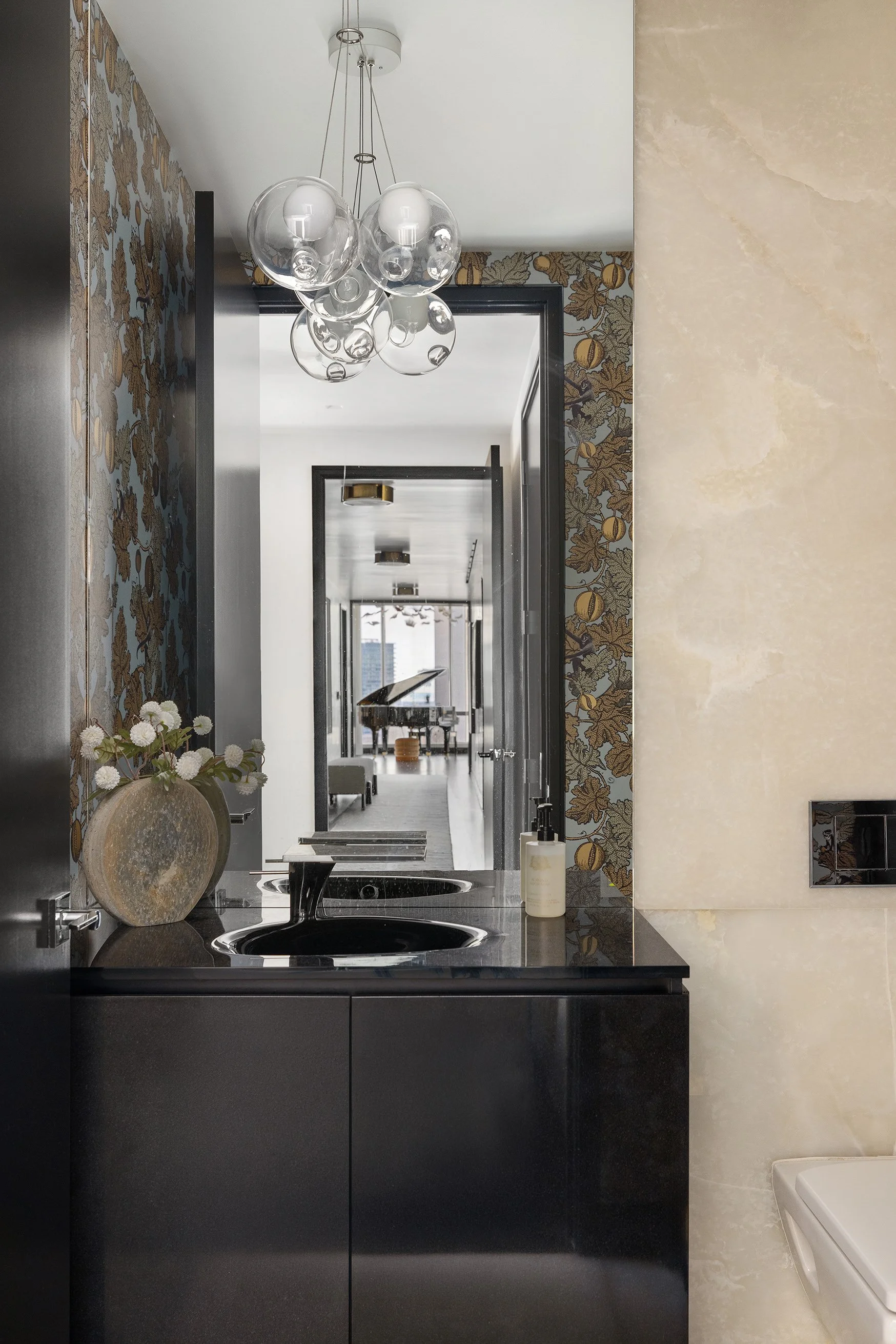View of a modern bathroom with a black double sink vanity, a large beige marble wall, and a decorative wallpapered wall with a pattern of acorns and leaves. A large glass bubble chandelier hangs from the ceiling, and a beige vase with flowers is on t