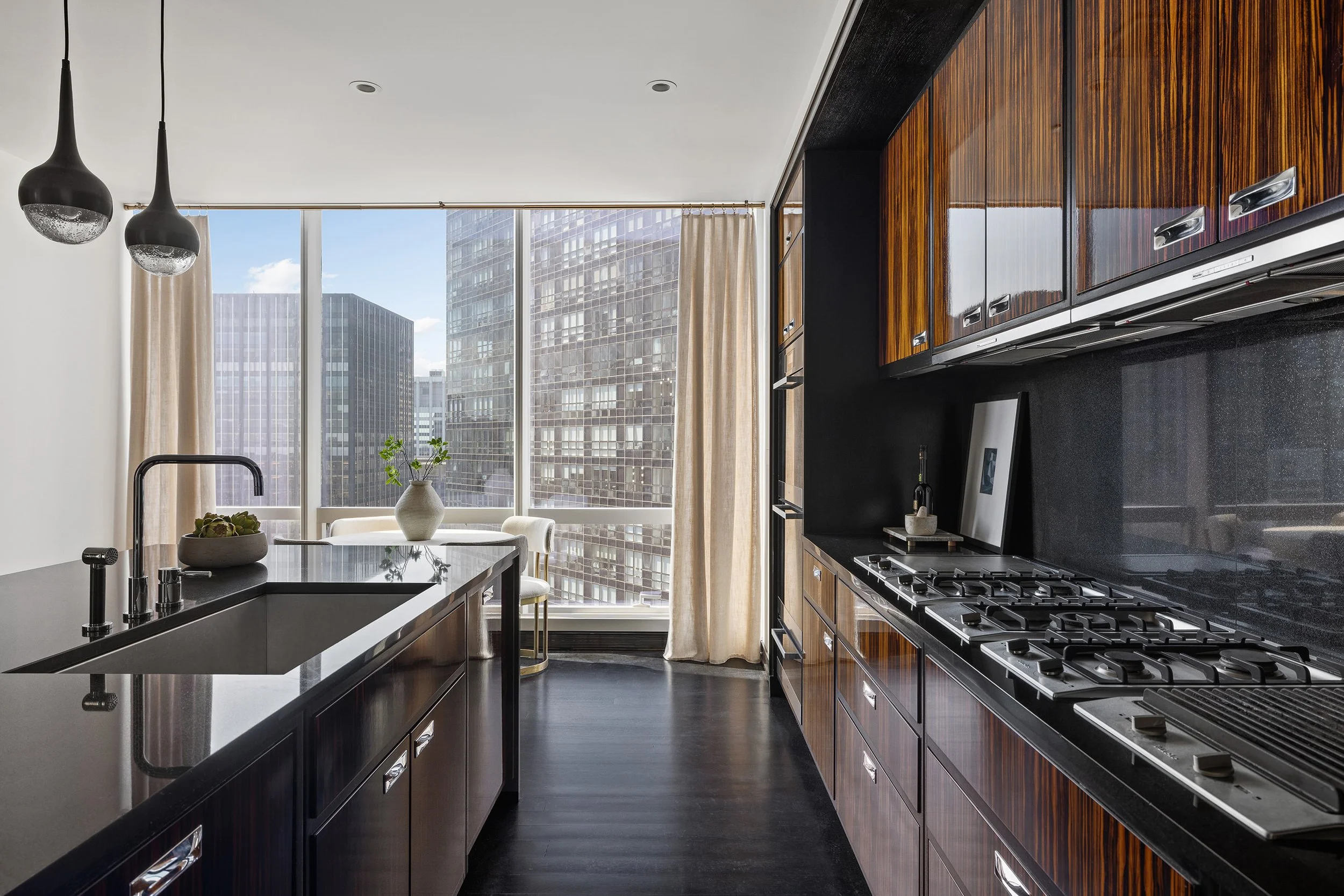 A modern kitchen with dark cabinetry, a black countertop, and a gas stove. Large windows with cityscape views, beige curtains, and hanging pendant lights.