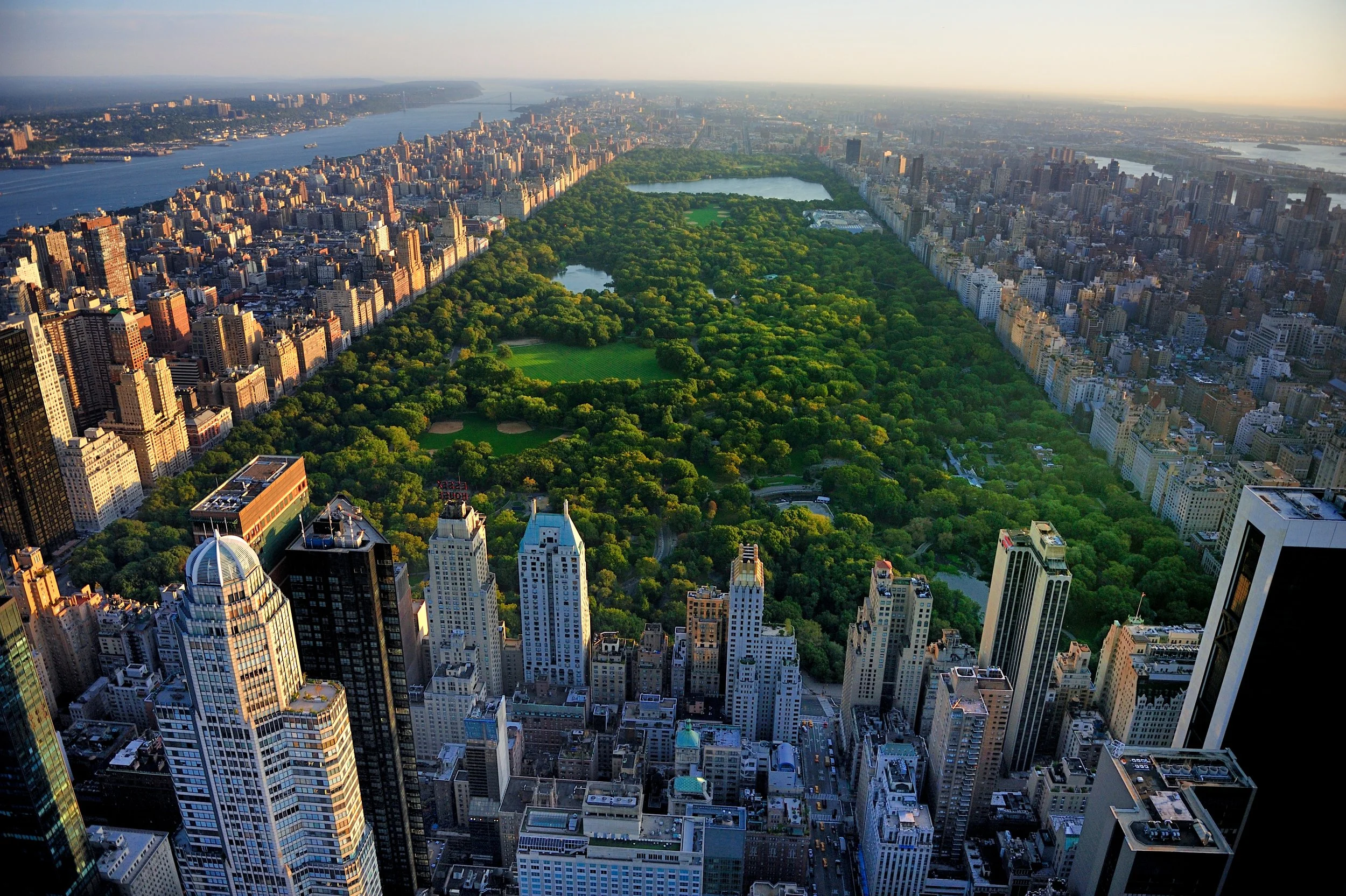 Aerial view of Central Park and surrounding New York City skyscrapers, with the city skyline and a body of water in the background.